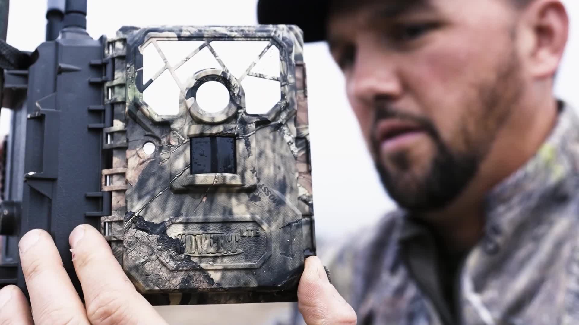 A man in camouflage holds a trail camera, its mottled pattern blending with the natural surroundings. He's studying the device intently, likely checking for activity captured on Wild TV.

