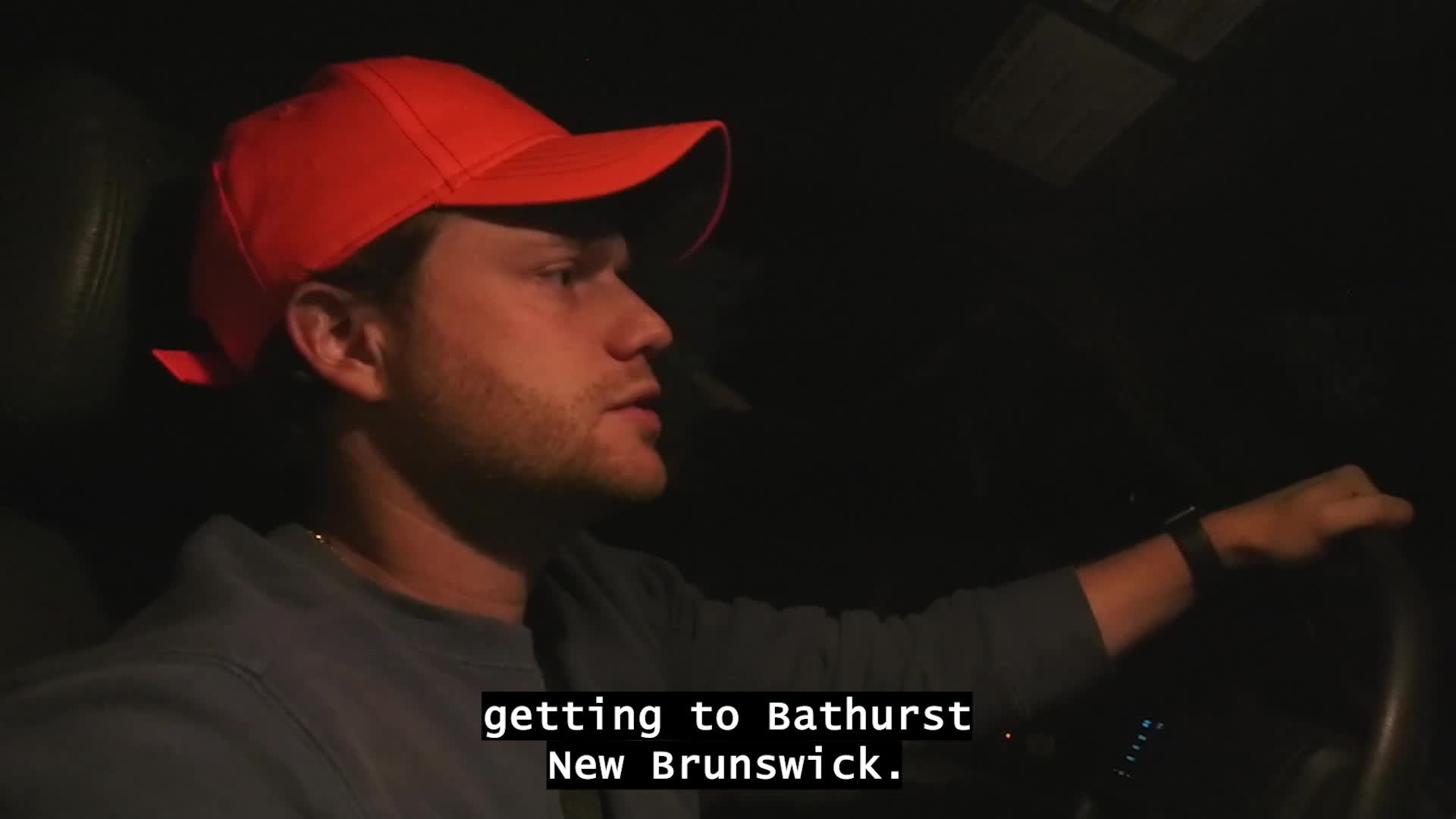 A man in an orange cap is driving, his face lit by the interior light. The caption on screen says they're headed to Bathurst, New Brunswick.
