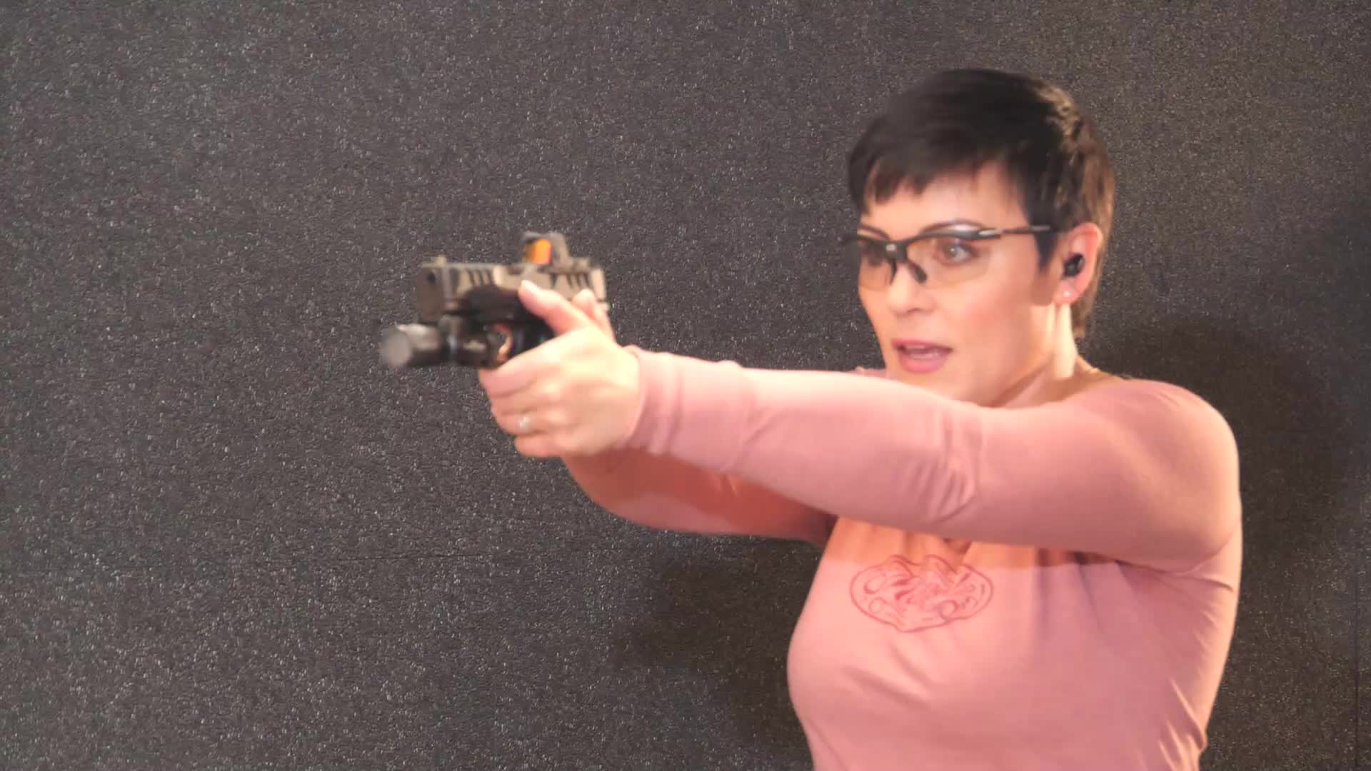 The woman, wearing safety glasses and ear protection, holds a pistol with both hands, aiming it forward. Her mouth is open as if she's about to speak or has just fired.
