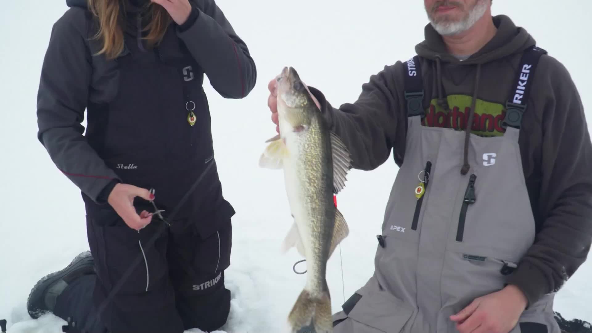 A man, kneeling on the ice, holds up a walleye, its silvery scales reflecting the overcast light. Beside him, a woman in dark overalls prepares to cut the line.
