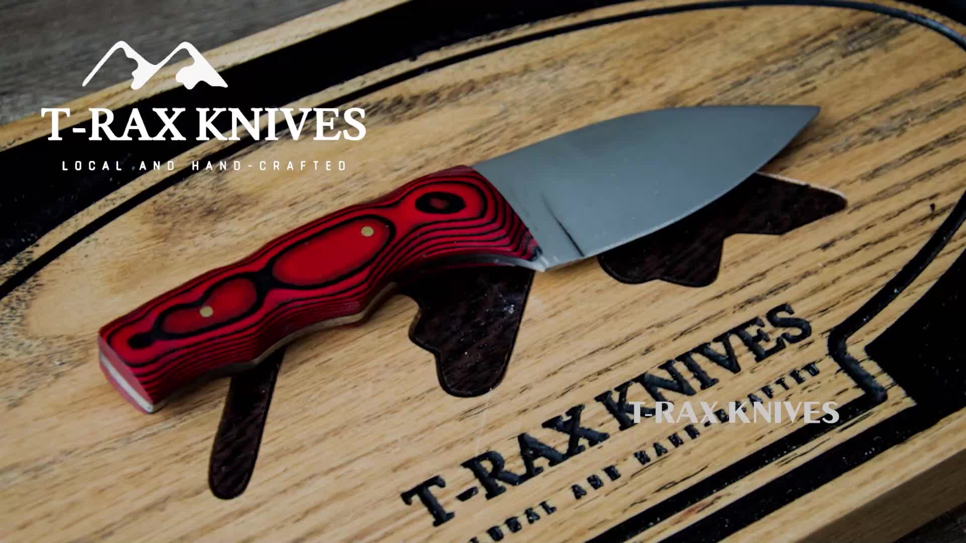 A handcrafted knife with a red and black handle rests on a wooden surface branded with the T-RAX Knives logo. The blade is polished silver, catching the light.
