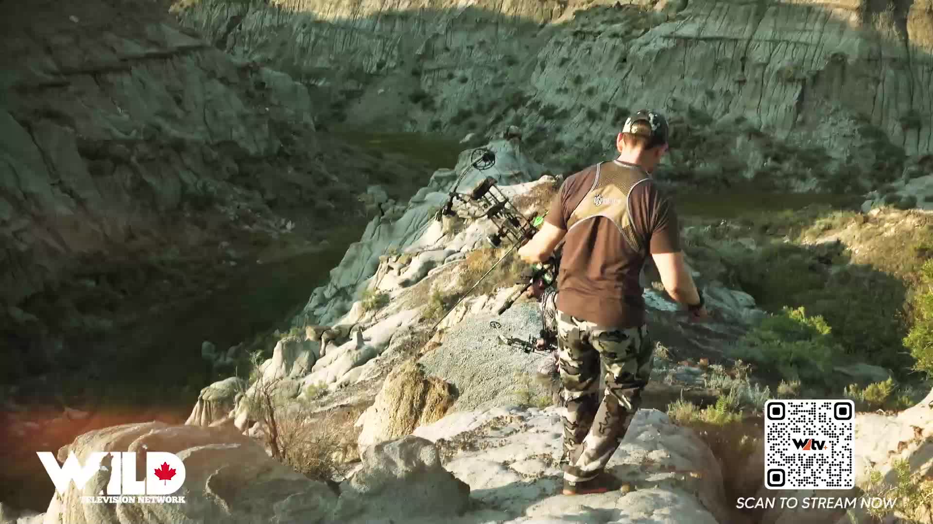 A man in camouflage pants and a brown shirt carefully walks across a rocky ridge, bow in hand. The landscape drops away dramatically behind him, a scene that could easily be on Wild TV.
