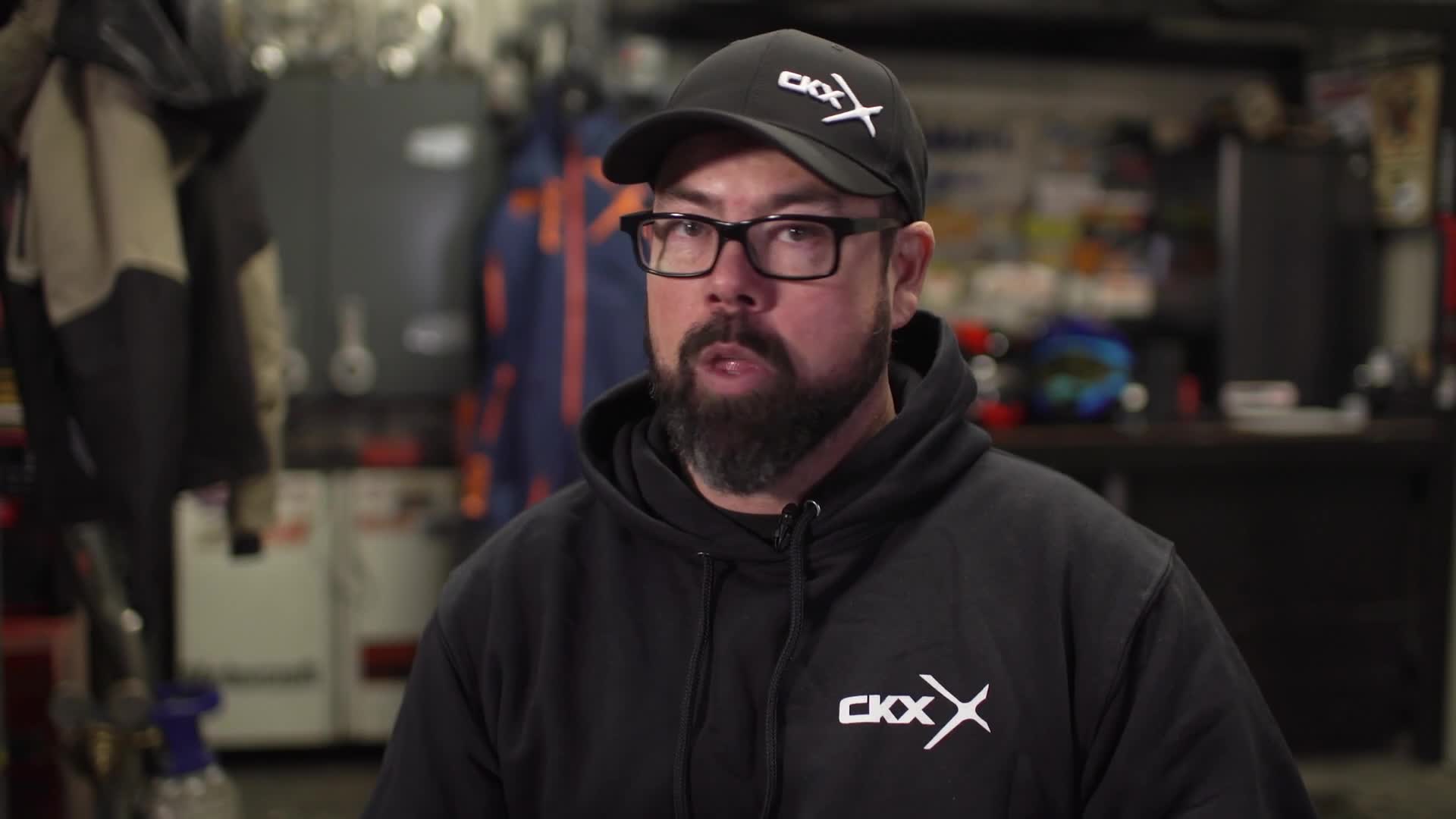 A man in a black cap and hoodie is talking directly to the camera, his lips moving as he speaks. Behind him, a cluttered workshop suggests a Canadian setting, perhaps for a Wild TV segment.
