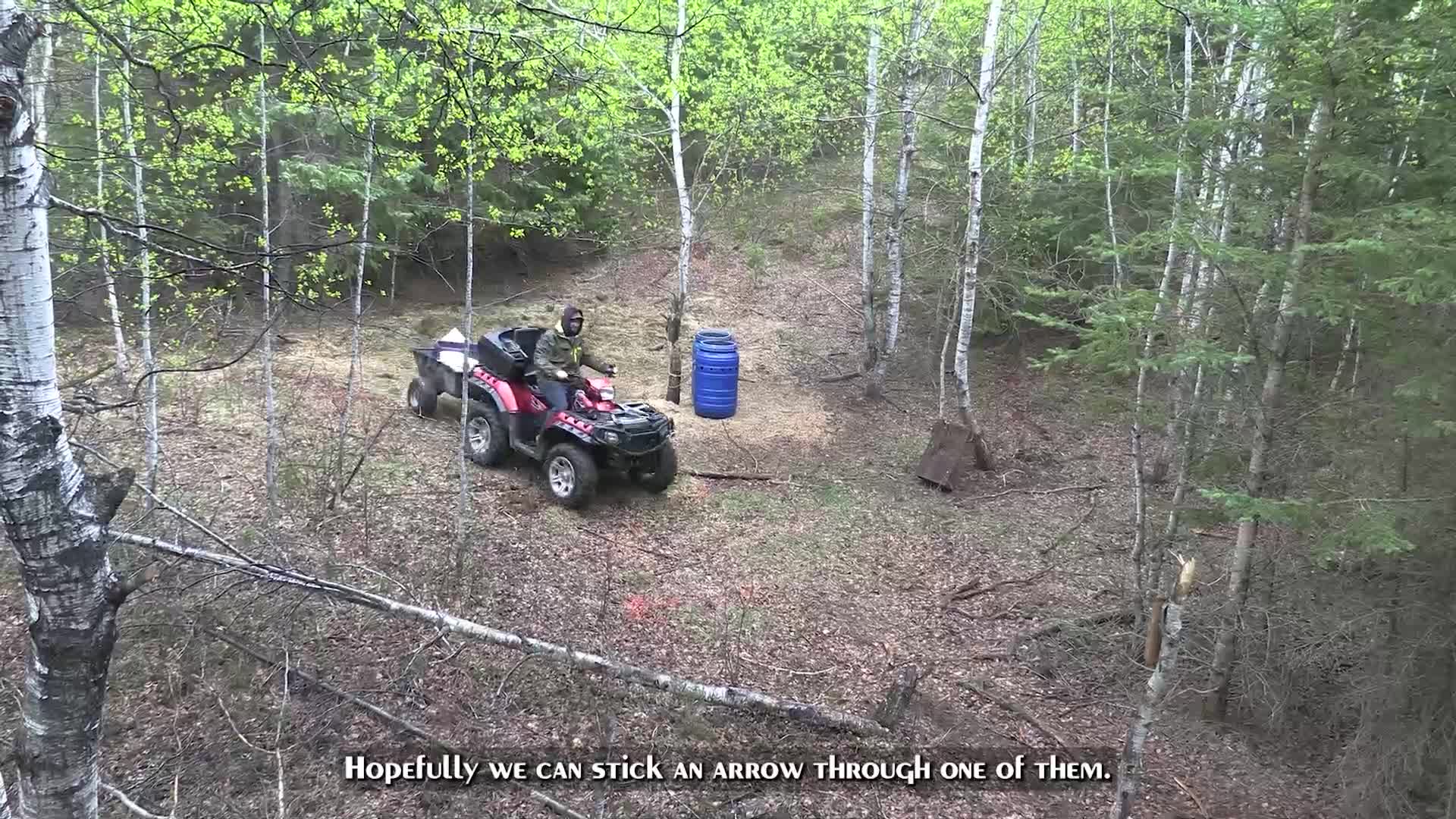 A red ATV with a trailer is parked on a wooded trail. A blue barrel sits nearby, and the trees are covered in fresh spring leaves. A red ATV with a trailer is parked on a wooded trail. A blue barrel sits nearby, and the trees are covered in fresh spring leaves.