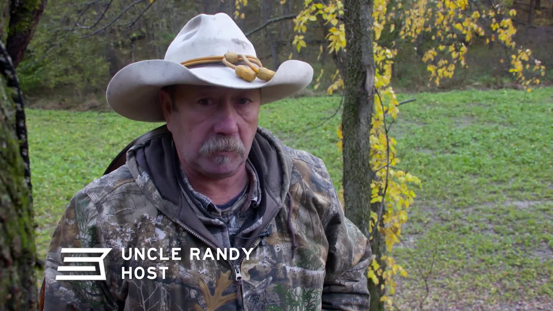 A man in a cowboy hat and camouflage jacket stands in a field of green grass, with yellowing leaves on trees behind him. He looks directly ahead, his expression serious. A man in a cowboy hat and camouflage jacket stands in a field of green grass, with yellowing leaves on trees behind him. He looks directly ahead, his expression serious.