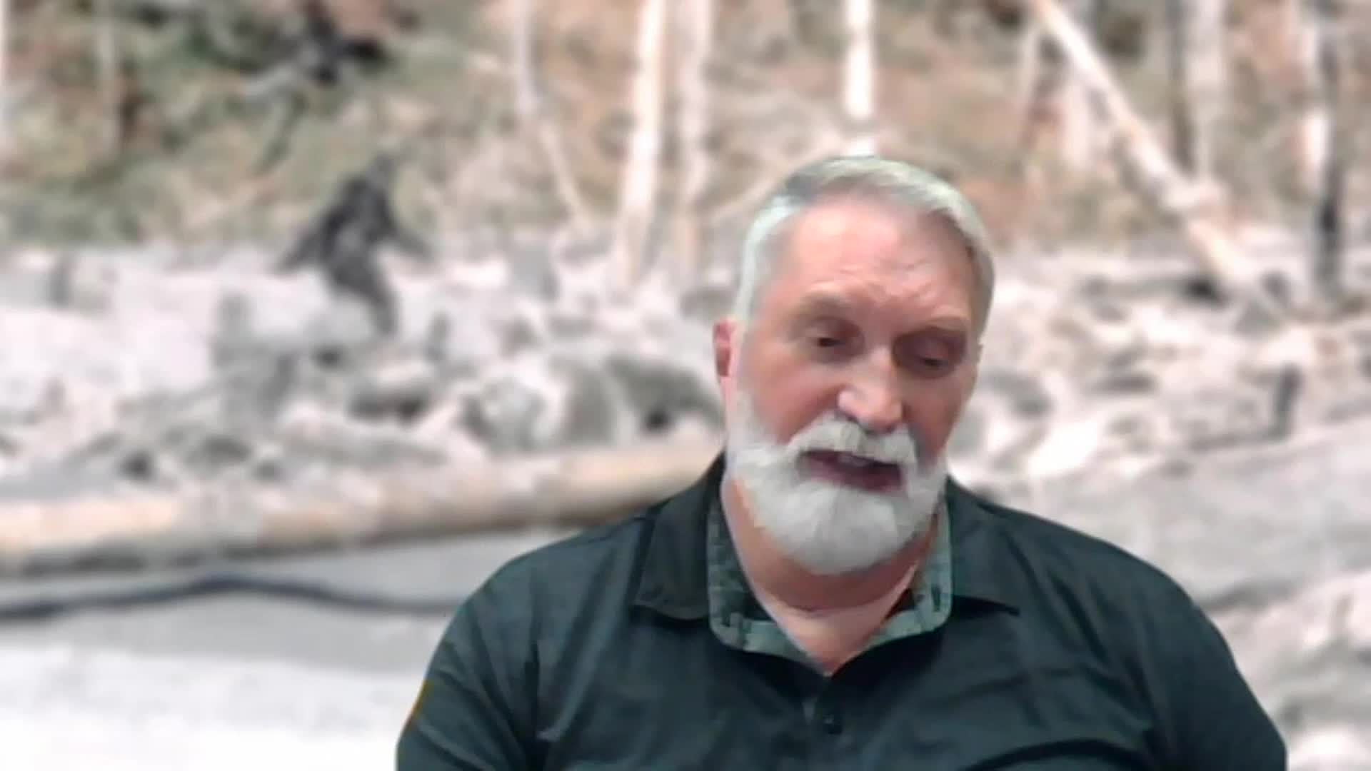 A man with a gray beard speaks, his expression earnest. Behind him, a shadowy figure stands amidst a blurry, wooded landscape, reminiscent of something you might see on Wild TV.