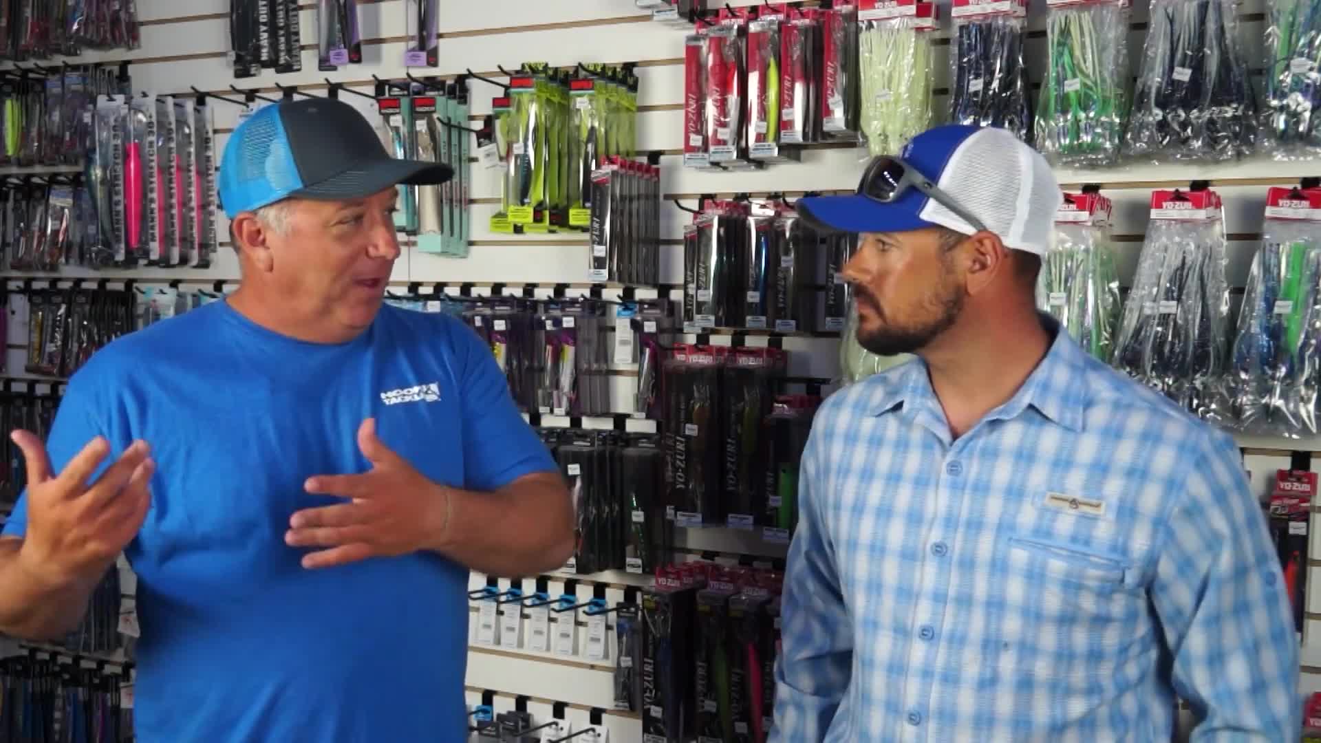 Two men stand in front of shelves stocked with fishing lures. The man on the left, wearing a blue shirt and a blue and grey trucker hat, gestures with his hands as he speaks. The man on the right, in a blue plaid shirt and a blue and white trucker hat, listens intently.