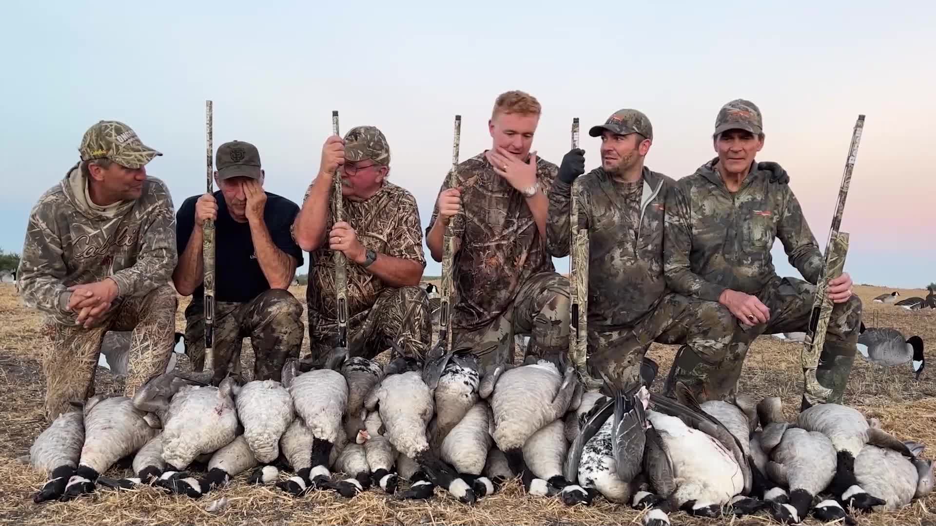 The group poses with their shotguns and a pile of harvested geese. The sky behind them is a pale, dusky blue.