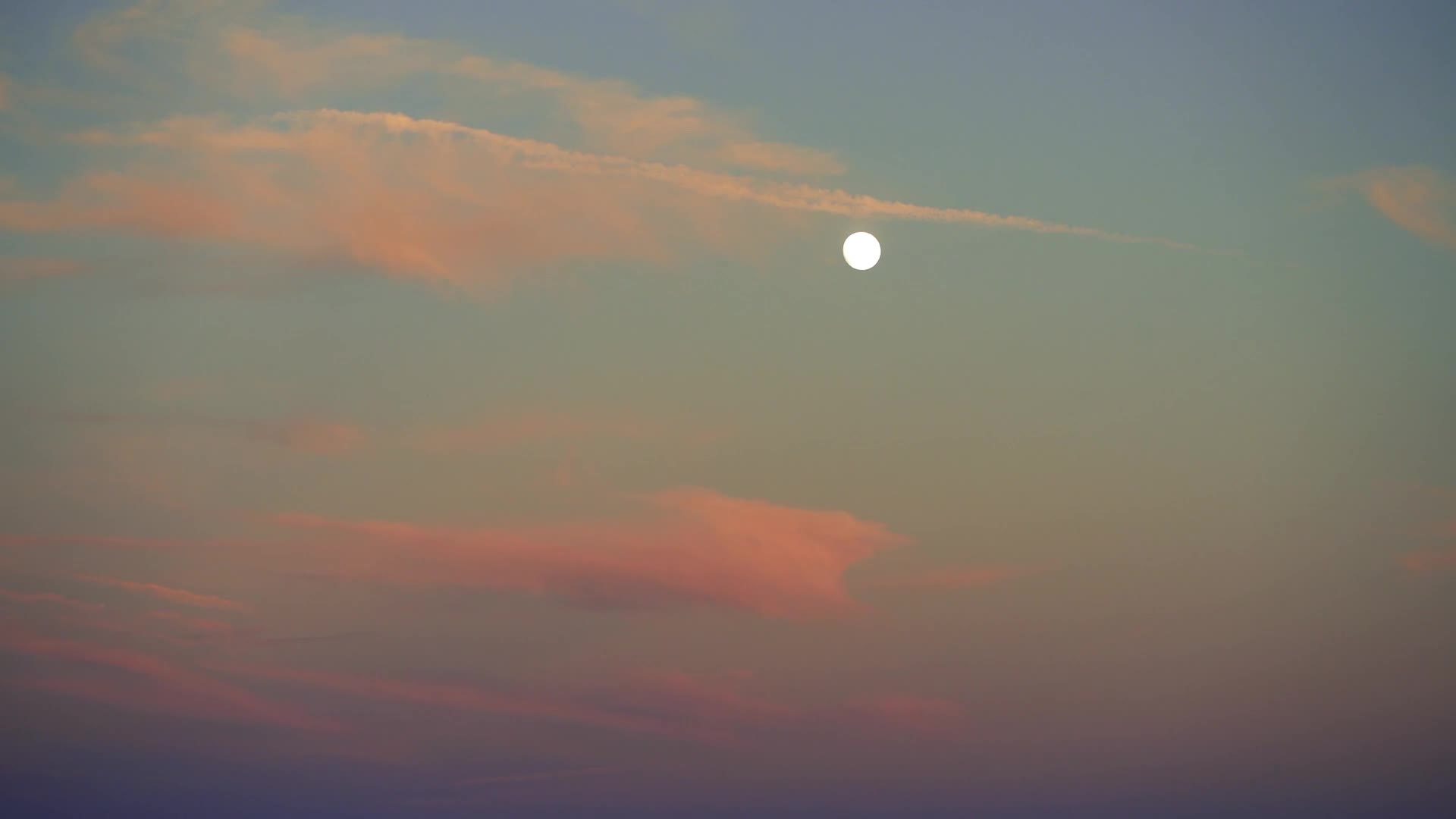 The moon hangs bright and round in a sky painted with soft pink and blue clouds. A thin, wispy cloud streaks across the horizon, catching the last light of day.