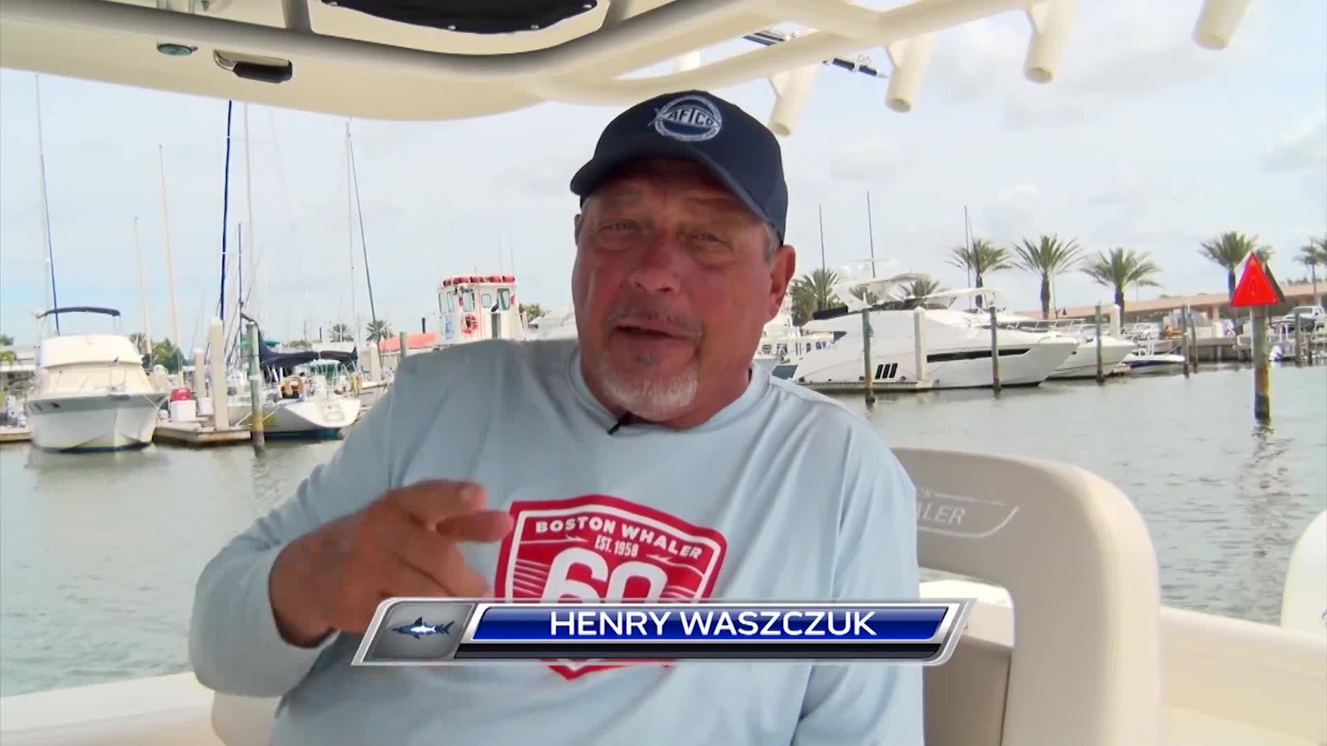 Henry Waszczuk, a familiar face from Wild TV, gestures while speaking from his boat. The marina behind him is filled with docked vessels under a bright, cloudy sky.