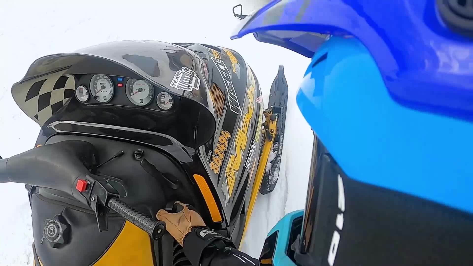 My gloved hand grips the handlebar of this snowmobile, its black and yellow decals sharp against the white snow. Another machine, bright blue, sits close beside me, ready for the ride.
