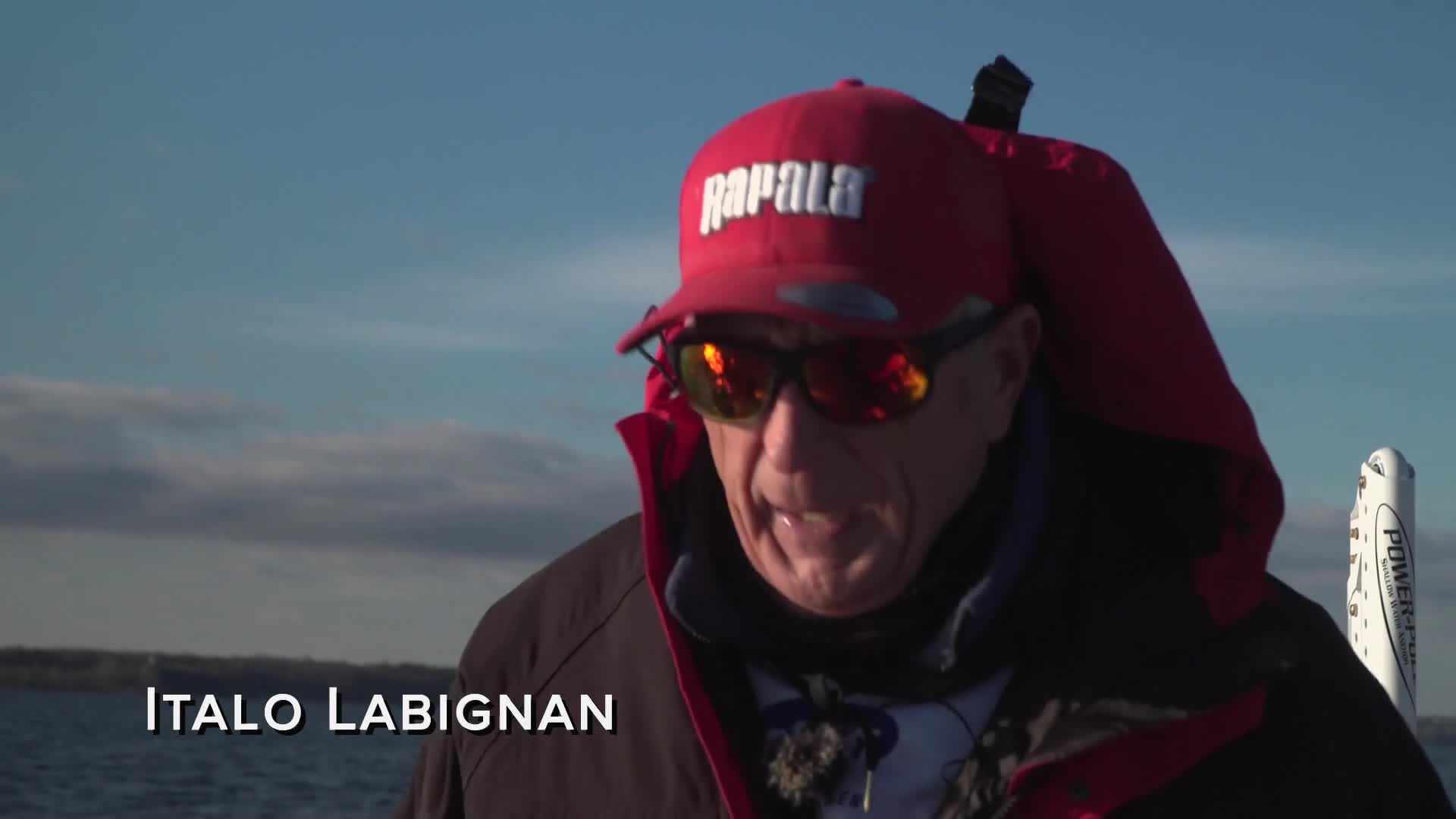 Italo Labignan, wearing a red Rapala hat, speaks into a microphone. A white Power-Pole is visible behind him against a bright blue sky.