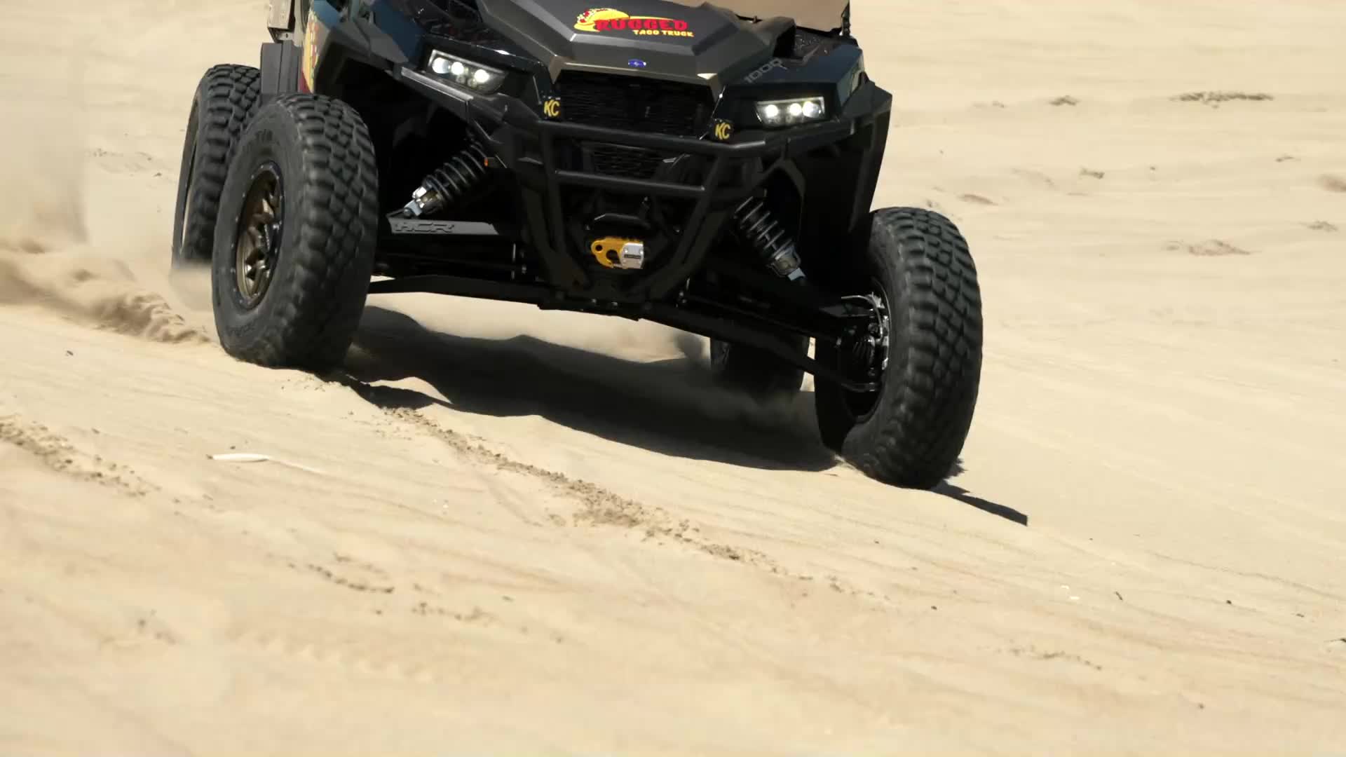 The side-by-side kicks up a spray of sand as it powers through the dunes. Its aggressive tires dig into the loose ground, propelling it forward.
