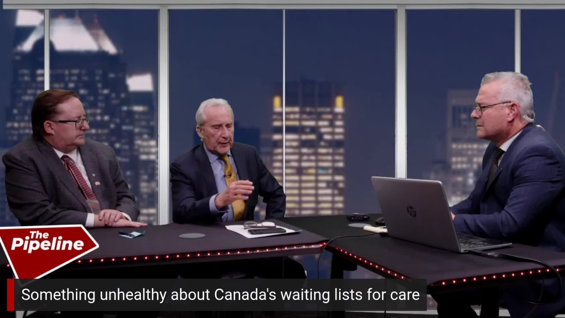Three men sit at desks in front of a city skyline at night. One man gestures with his hand while speaking. A laptop sits open on the desk of the man to his right.
