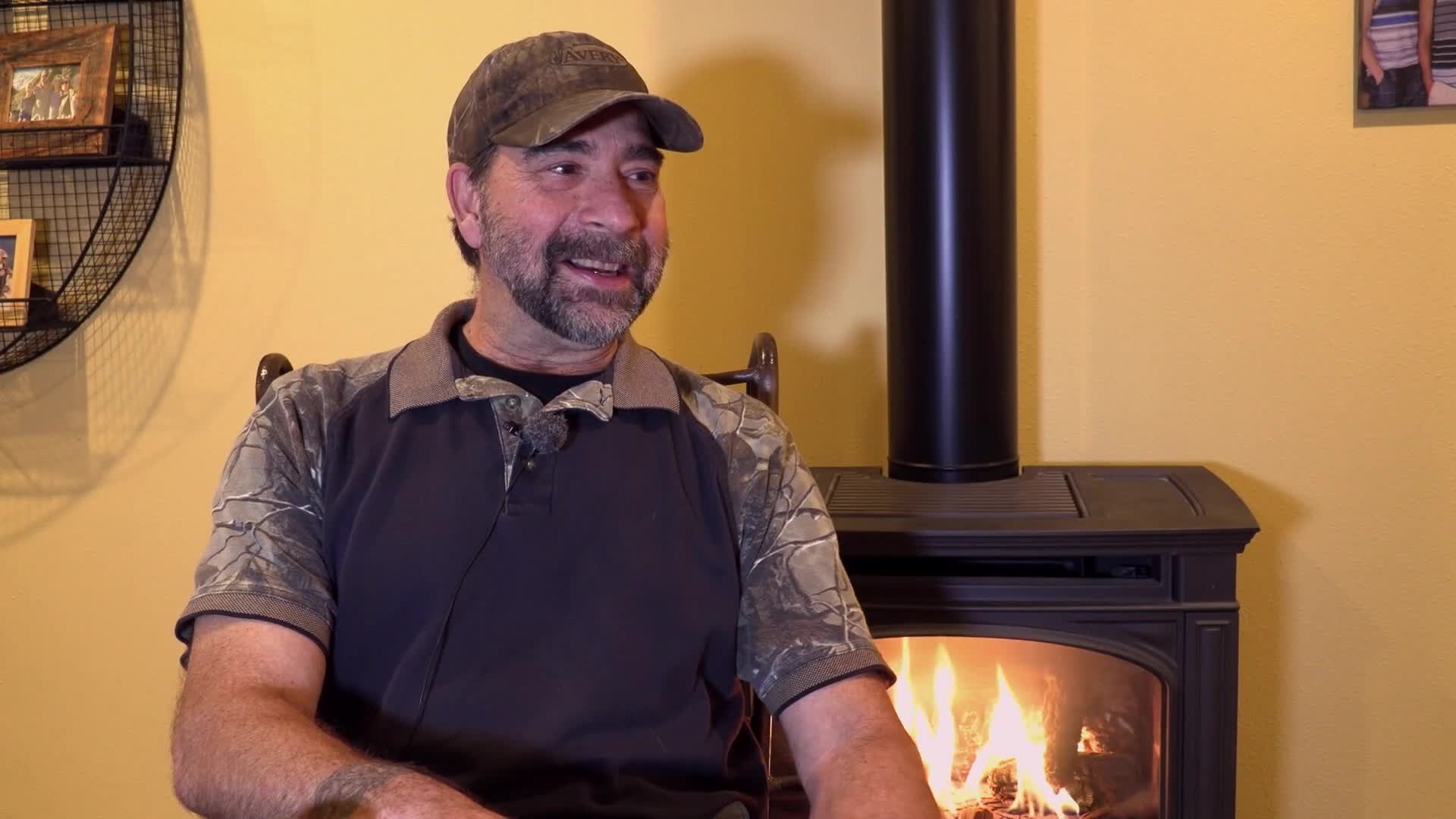 A man in a camo shirt and baseball cap speaks animatedly, a wood stove with a bright fire crackling behind him. The scene feels like a segment from Wild TV, perhaps a story from the Canadian outdoors.
