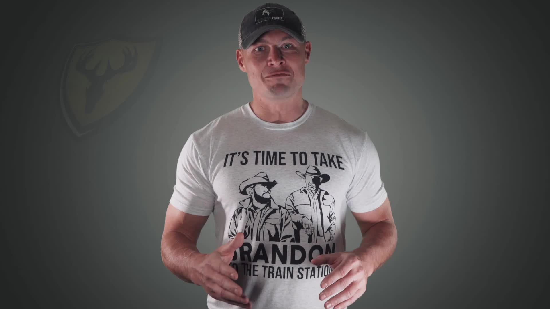 A man in a grey t-shirt stands before a dark backdrop. His shirt features a graphic of two men in cowboy hats and the text "IT'S TIME TO TAKE BRANDON TO THE TRAIN STATION." A man in a grey t-shirt stands before a dark backdrop. His shirt features a graphic of two men in cowboy hats and the text "IT'S TIME TO TAKE BRANDON TO THE TRAIN STATION."
