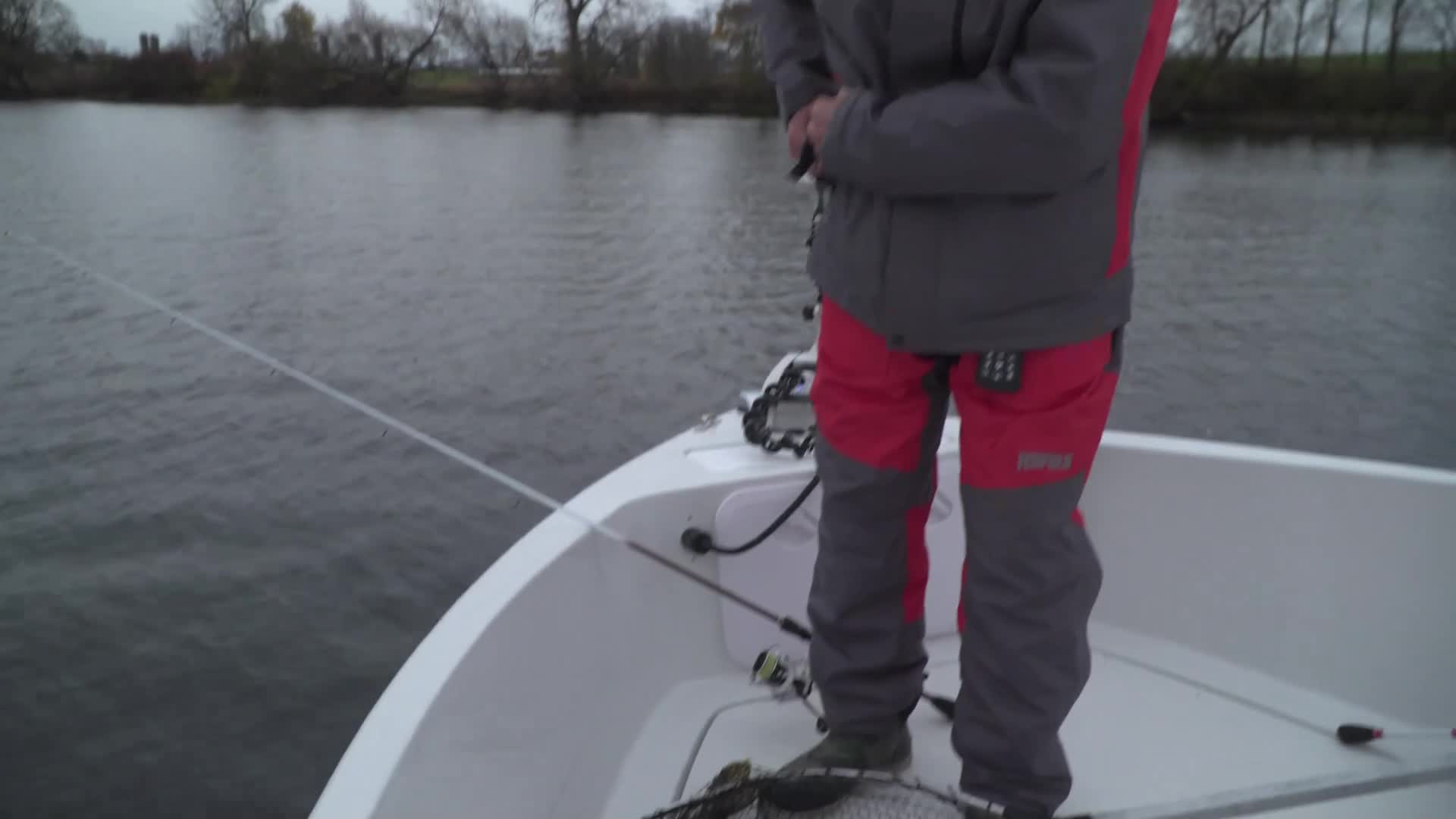 A person in a grey and red jacket and pants stands in a white boat, holding a fishing rod. The line stretches taut across the calm, grey water toward the distant, tree-lined shore. A person in a grey and red jacket and pants stands in a white boat, holding a fishing rod. The line stretches taut across the calm, grey water toward the distant, tree-lined shore.