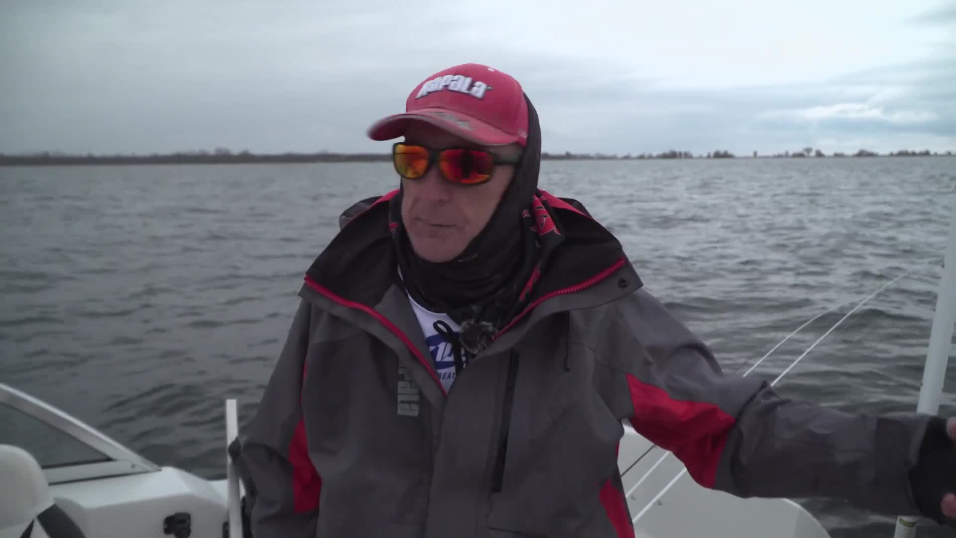 The boat cuts through the choppy grey water under an overcast sky. A man wearing a red Rapala hat and sunglasses looks out at the horizon. The boat cuts through the choppy grey water under an overcast sky. A man wearing a red Rapala hat and sunglasses looks out at the horizon.