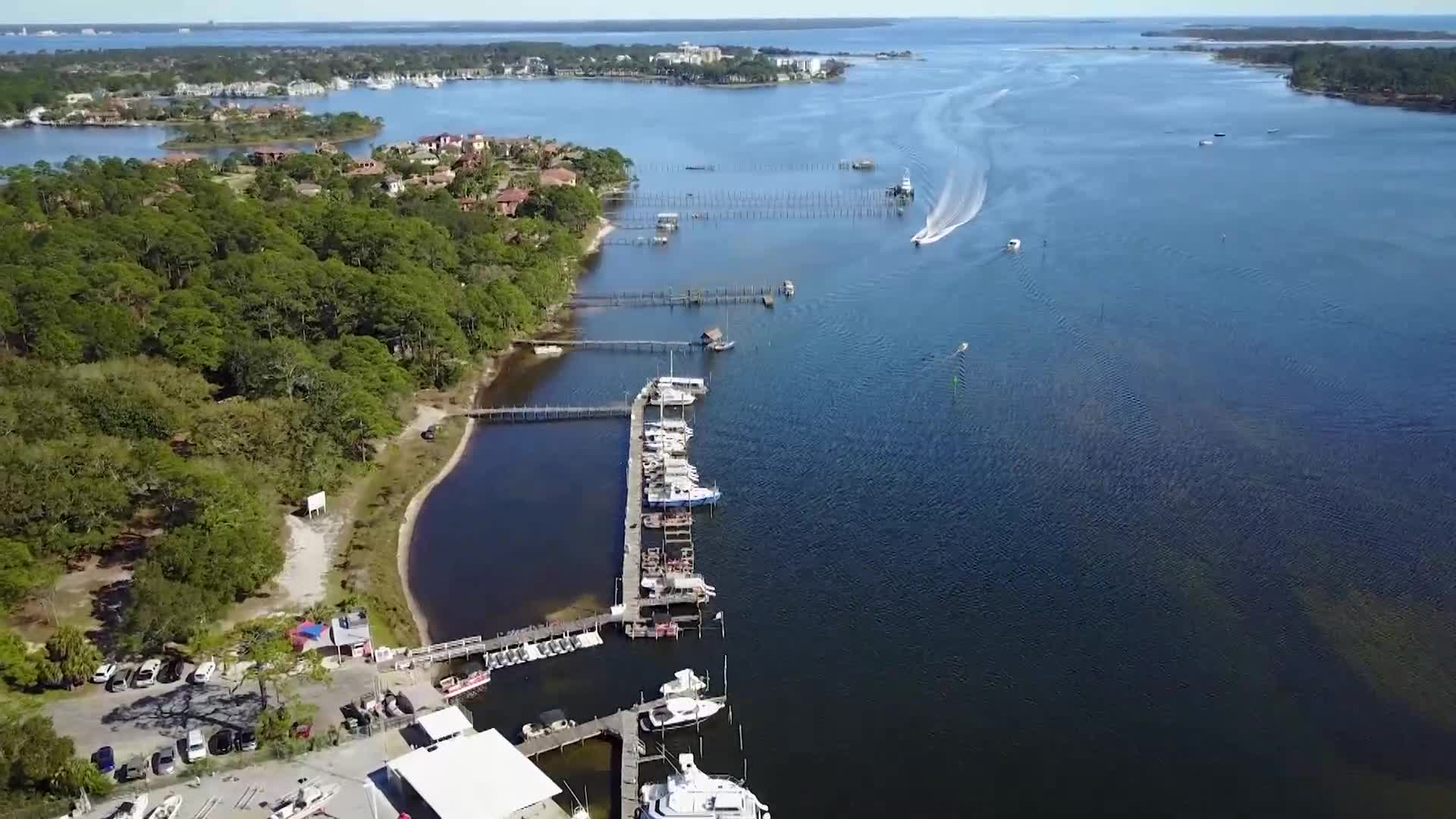 A white boat speeds away, leaving a frothy wake on the deep blue water. Several other boats are moored at a long wooden dock, nestled against a tree-lined shore. A white boat speeds away, leaving a frothy wake on the deep blue water. Several other boats are moored at a long wooden dock, nestled against a tree-lined shore.