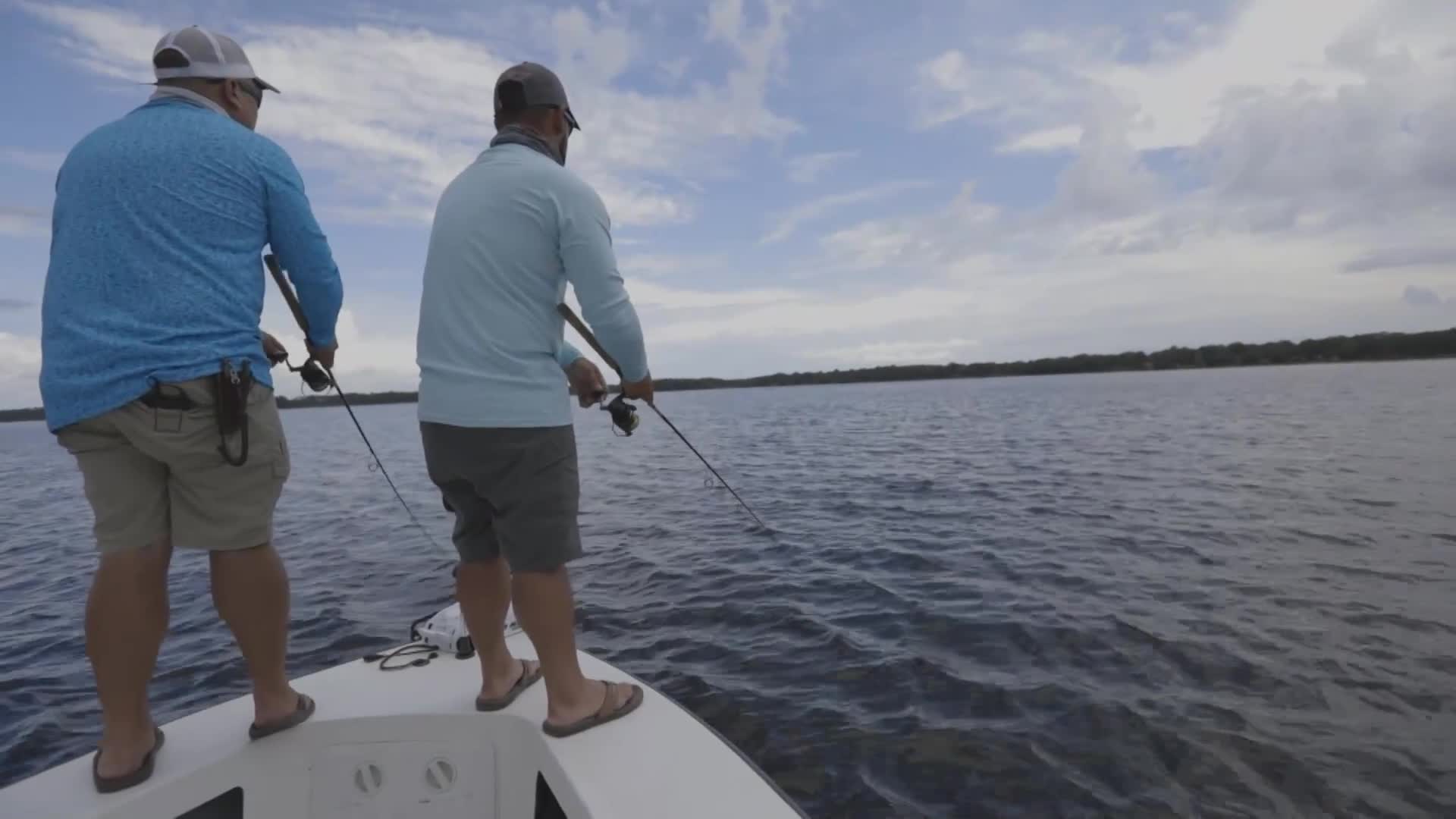 Two guys are casting their lines from the bow of a boat on a wide, calm lake. The sky above is a mix of blue and white clouds, promising a good day for fishing, perhaps for a segment on Wild TV.