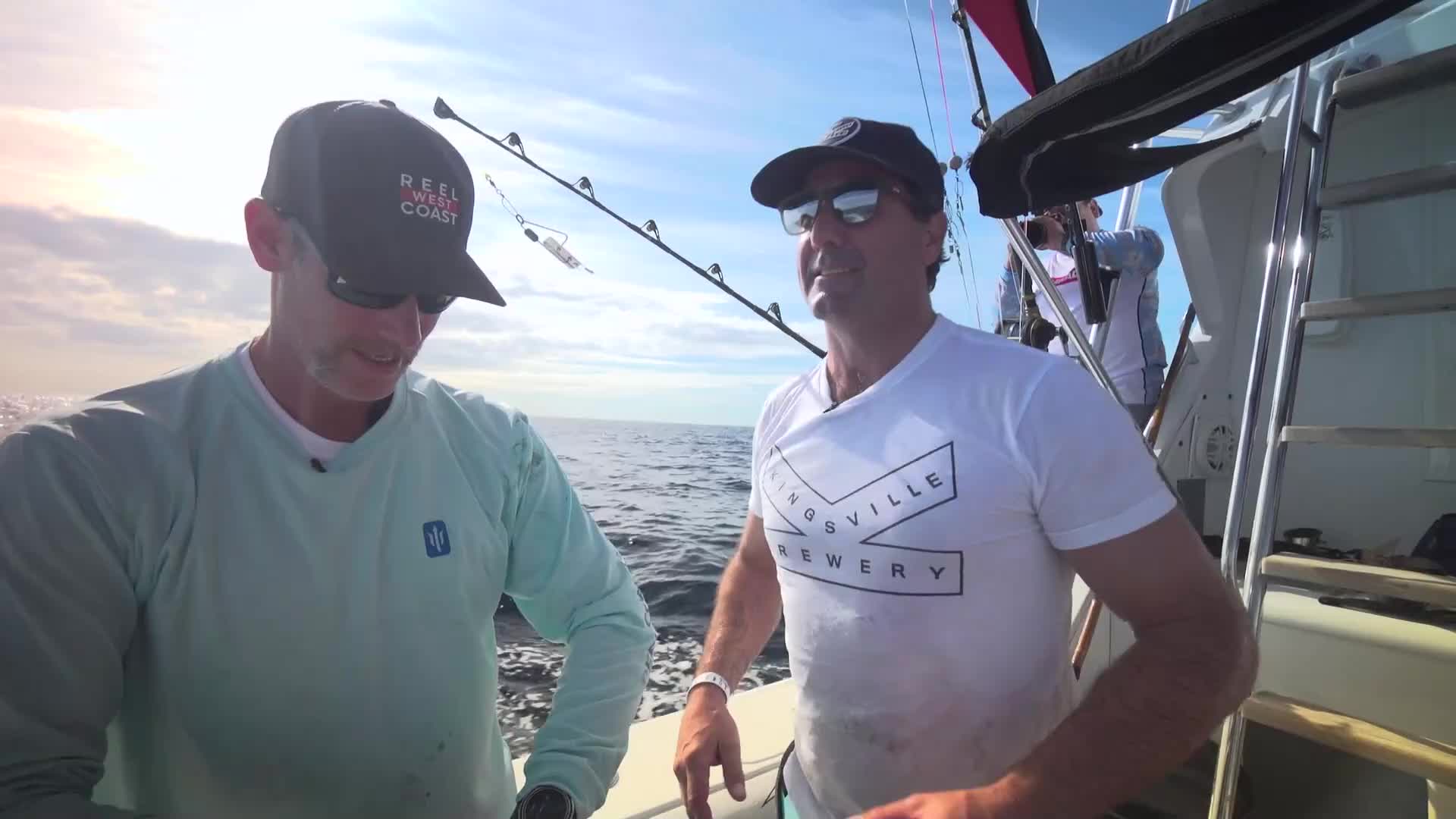 The sun glints off the ocean as two men stand on a boat, discussing their fishing expedition. One man, wearing a cap that reads "Reel West Coast," looks down at something in his hands.