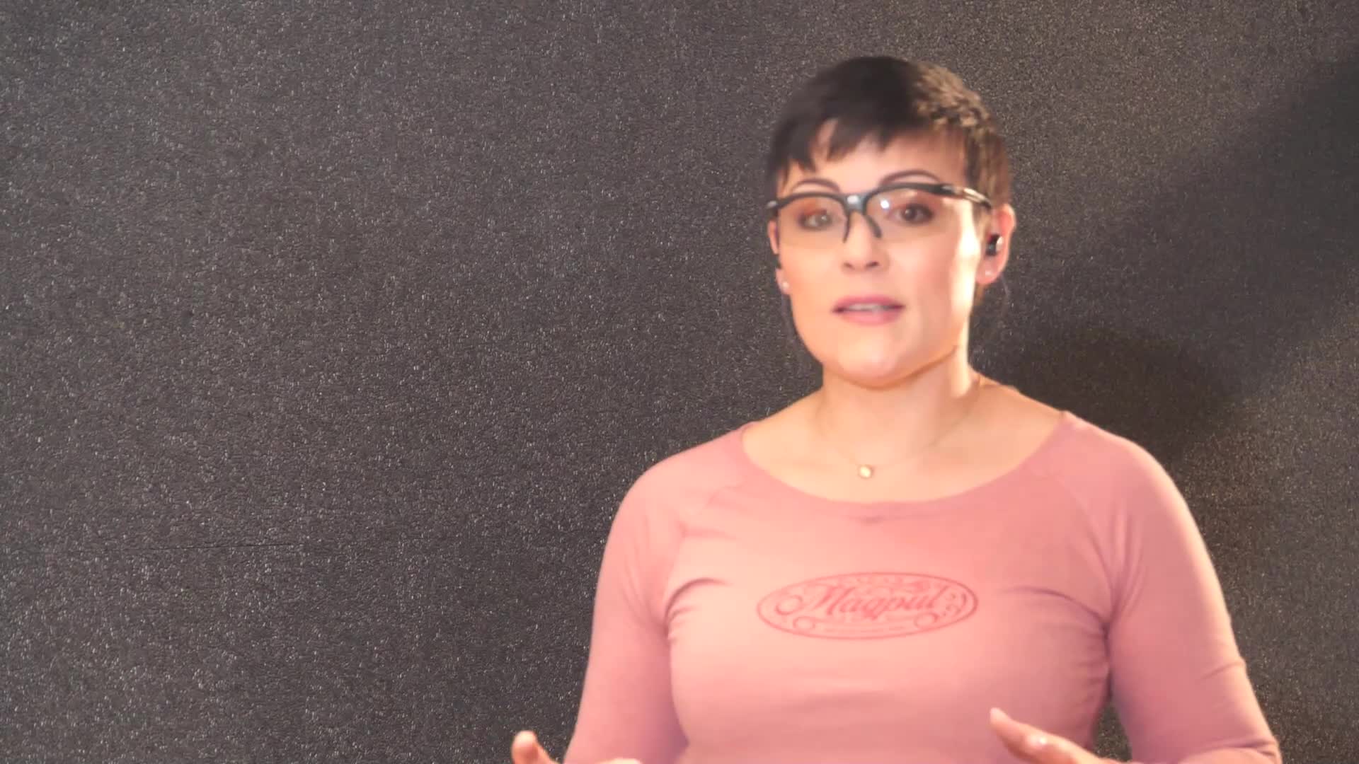 A woman with short dark hair and glasses is speaking, her hands gesturing slightly. She wears a rose-colored long-sleeved shirt with a logo on the chest.