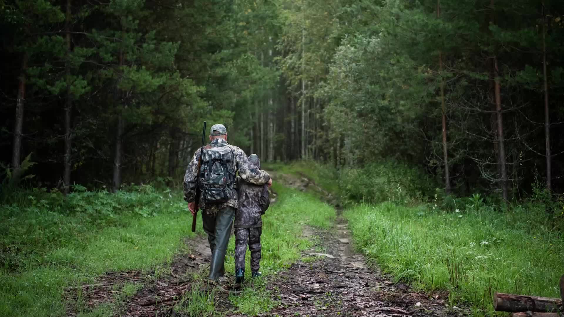 A man and a child, dressed in camouflage, walk down a muddy path into the woods. The man has a rifle slung over his shoulder, and his arm is around the child's shoulders.