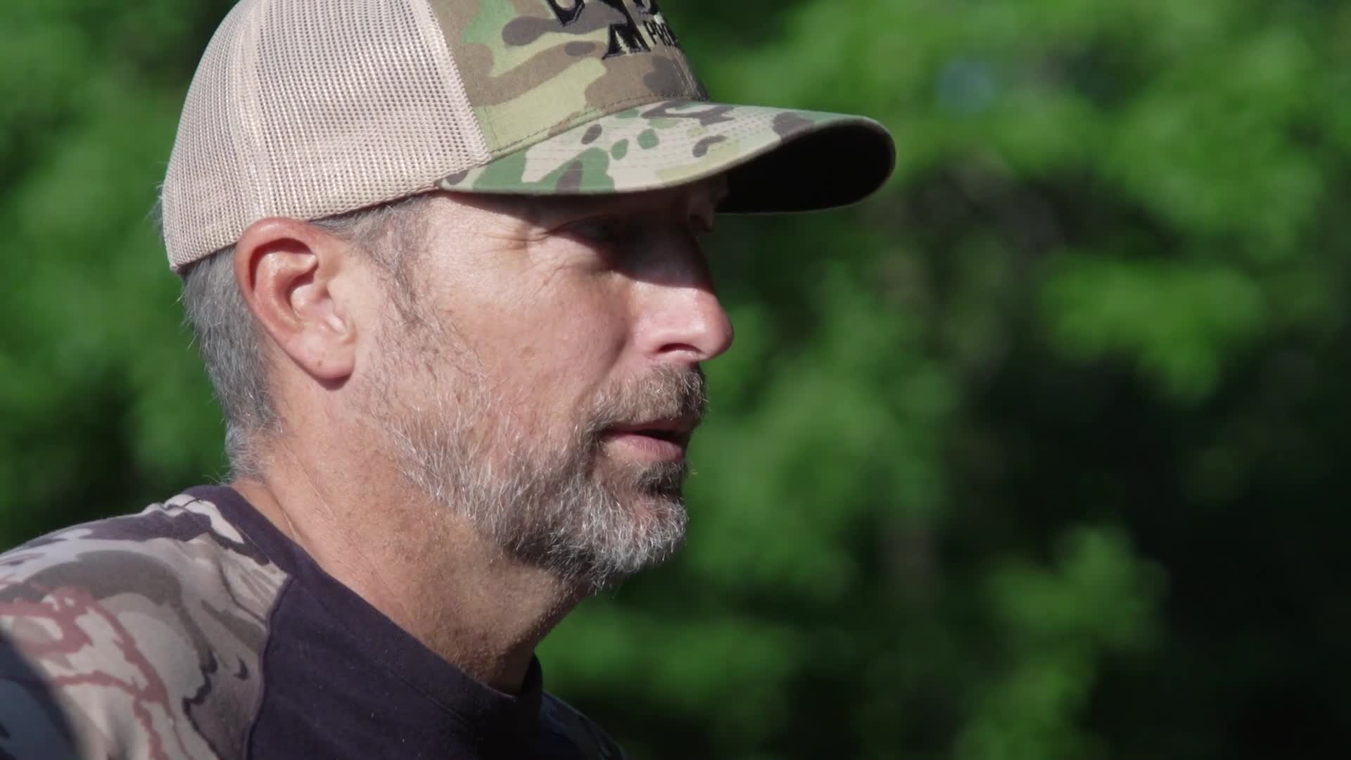The man in the camouflage hat turns his head, his gaze fixed on something beyond the frame. He's wearing a shirt with a similar pattern, blending into the backdrop of green trees.