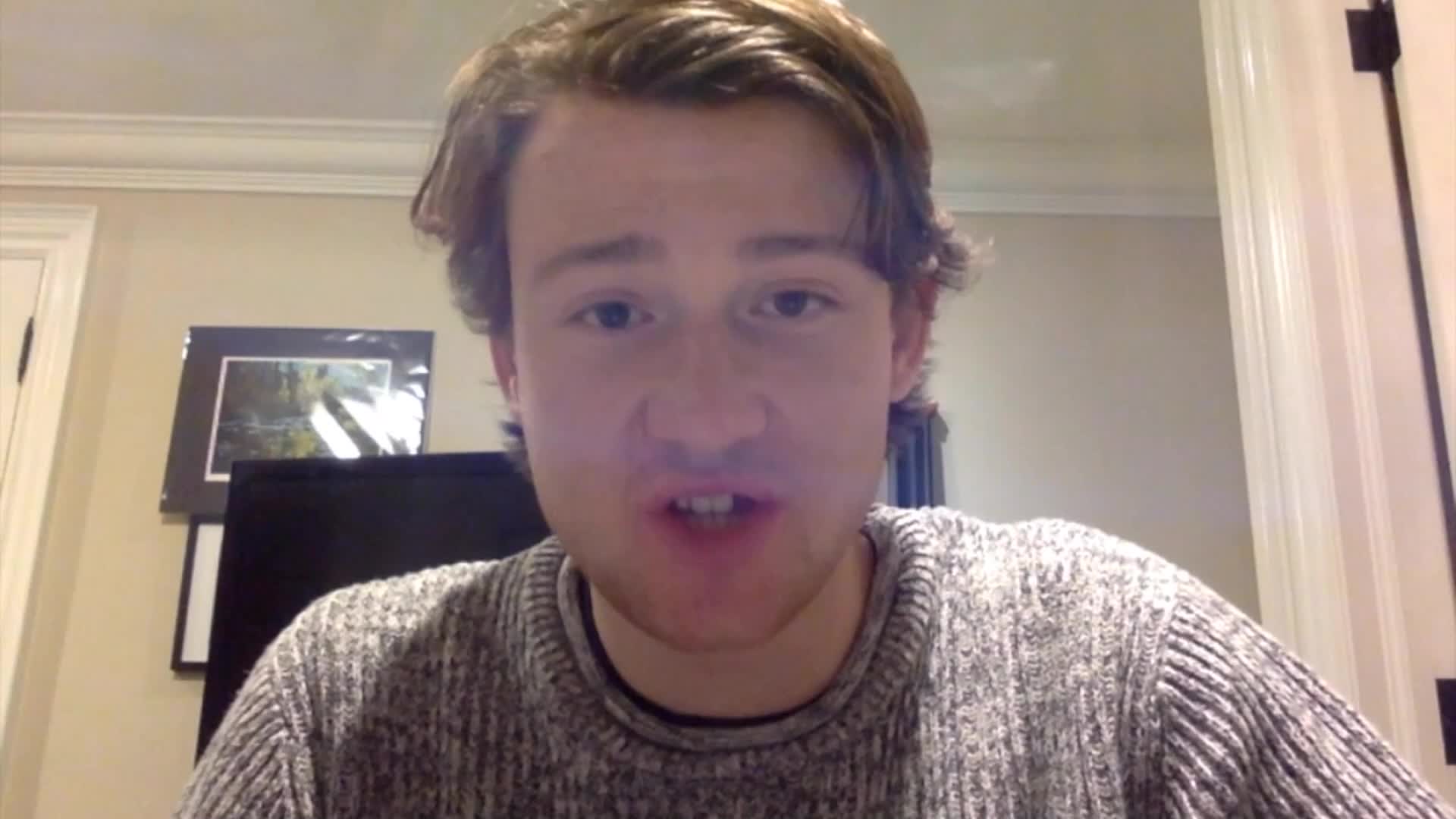 A young man with light brown hair, wearing a grey knitted sweater, speaks directly to the camera. Behind him, a framed picture hangs on the wall.