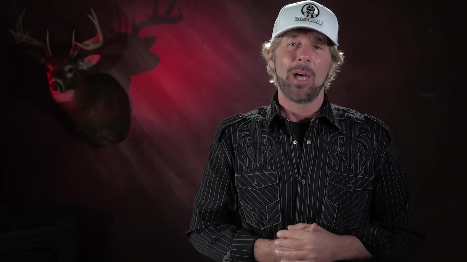 A man in a black pinstripe shirt and a white cap speaks into the camera. Behind him, a mounted deer head casts a shadow against a red-lit backdrop.