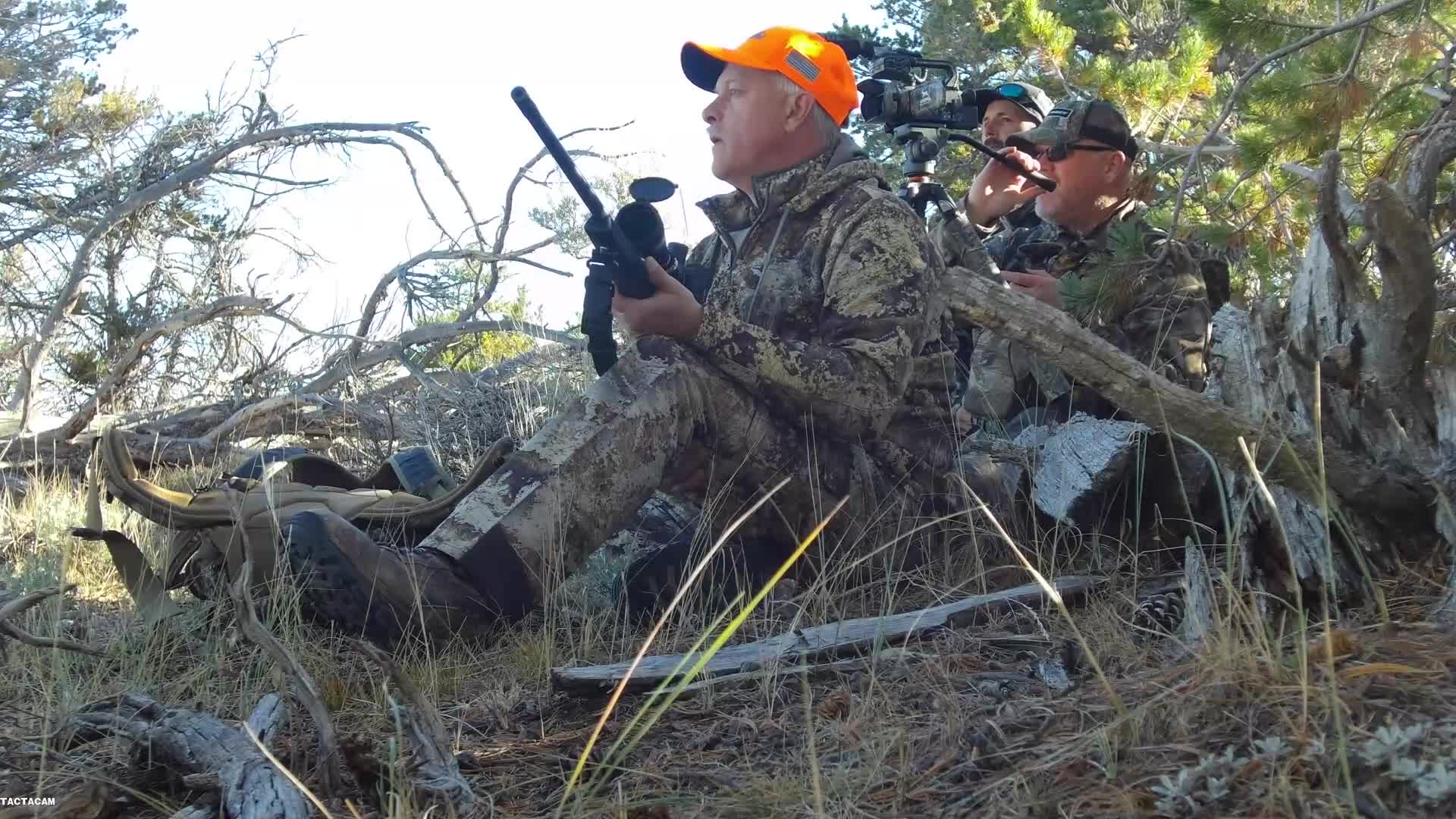 A man in an orange hat and camouflage gear holds a rifle, scanning the brush. Another man behind him, also in camouflage, operates a video camera on a tripod.