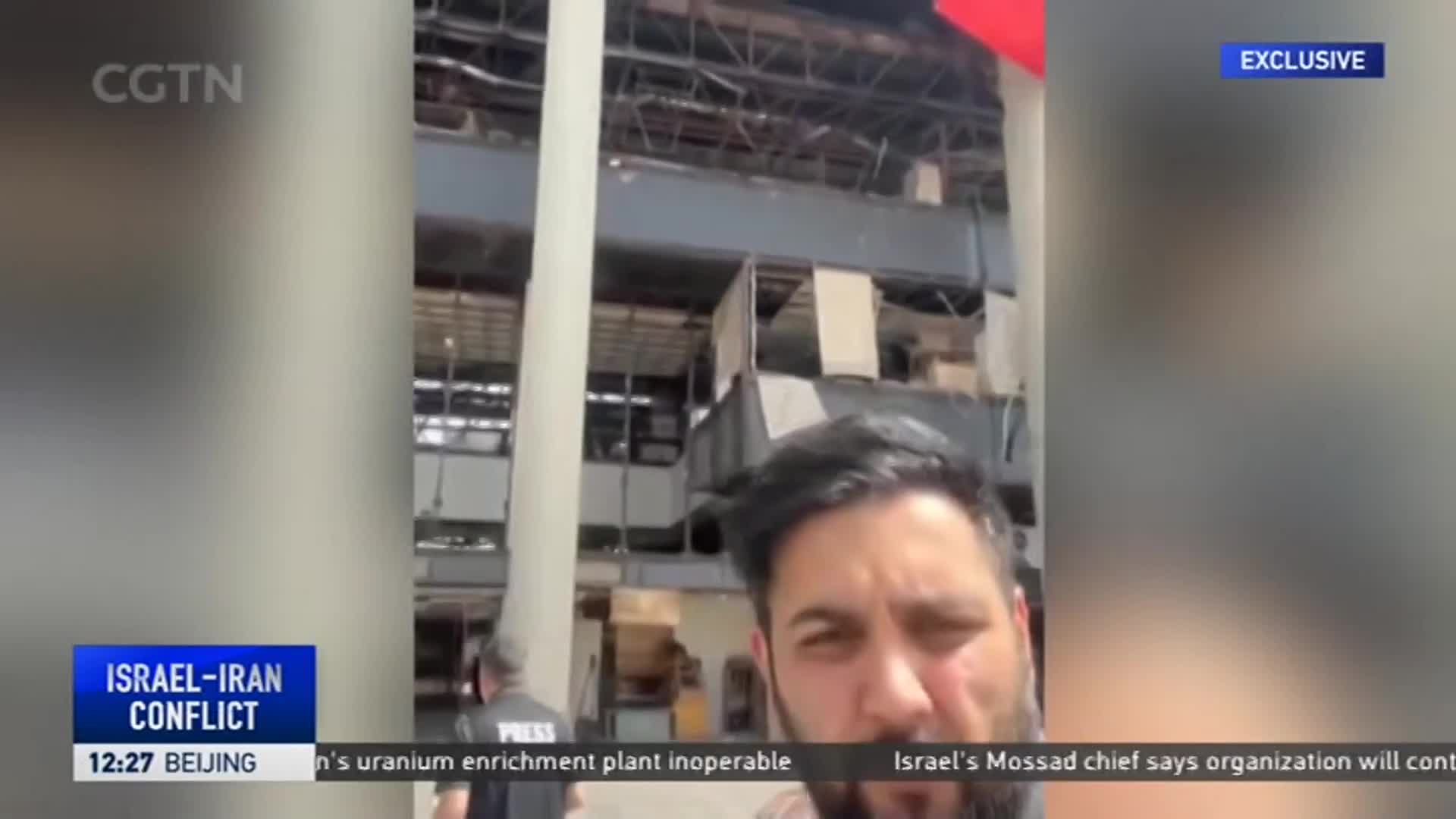 A man is filming the damaged facade of a building, likely for CGTN, with the "Israel-Iran Conflict" as the backdrop. The structure's upper levels show significant damage, with exposed beams and missing panels.
