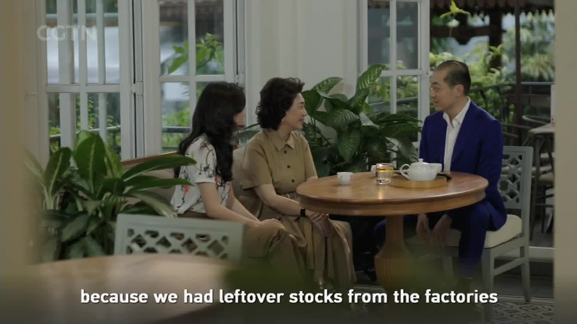 Three people sit around a small, round table, engaged in conversation. A man in a blue suit gestures as he speaks, while the two women listen attentively, a teapot and cups resting on the table.
