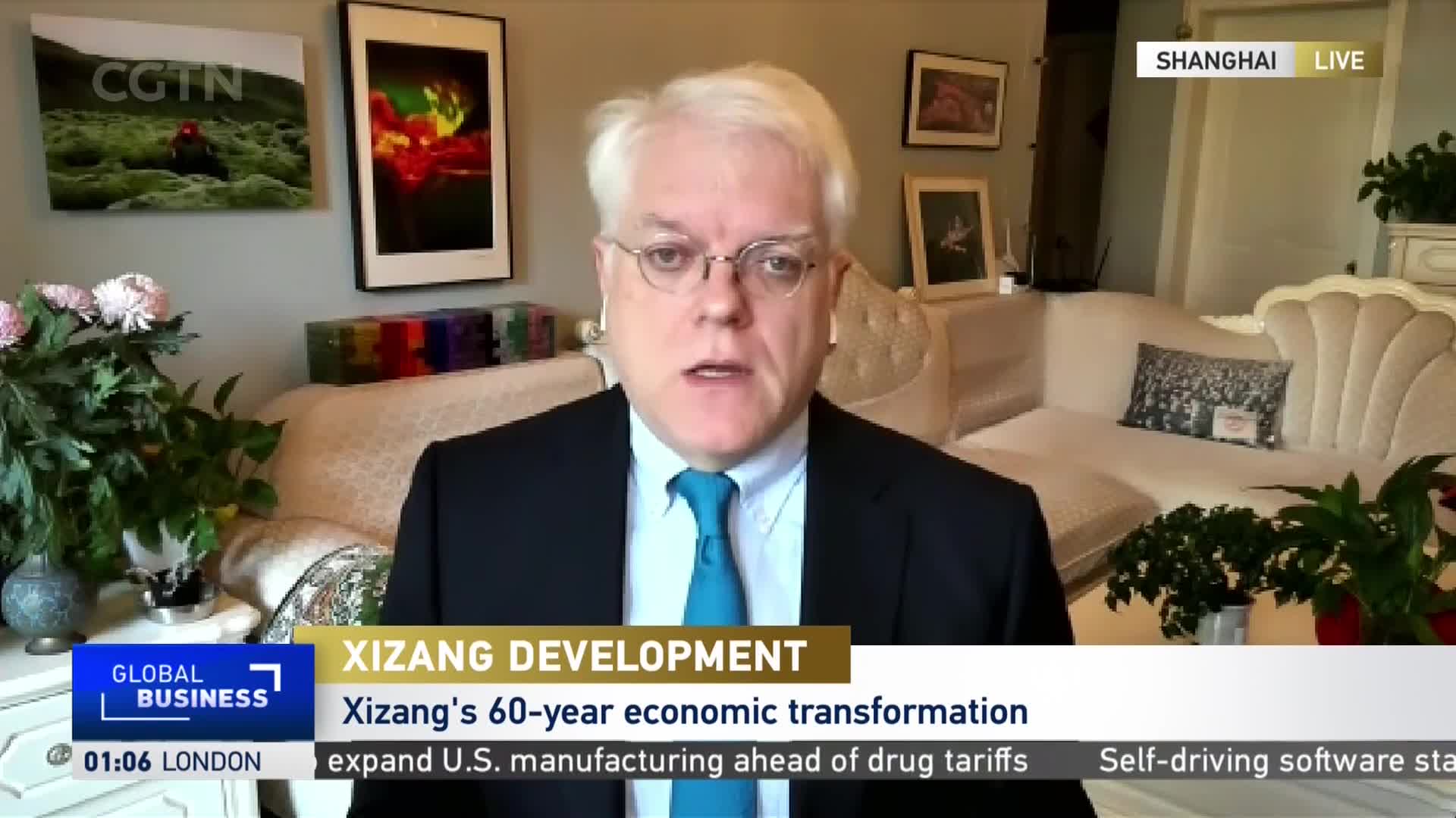 A man in a suit and tie speaks directly to the camera, framed by a couch and floral arrangements. The CGTN logo and a lower-third graphic about Xizang's economic transformation are visible on the screen.

