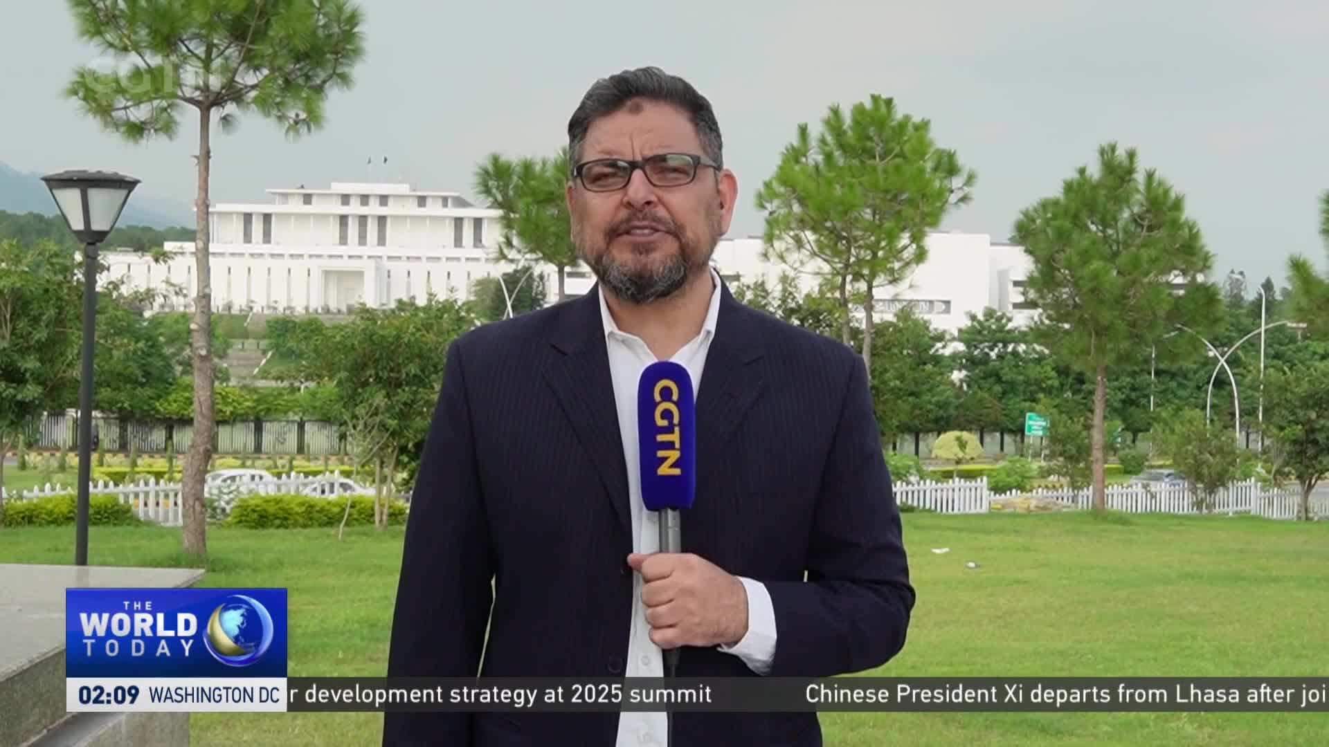A man in a dark suit, holding a CGTN microphone, speaks in front of a large white building. The scene is framed by green lawns and trees, with a news ticker running along the bottom of the screen.
