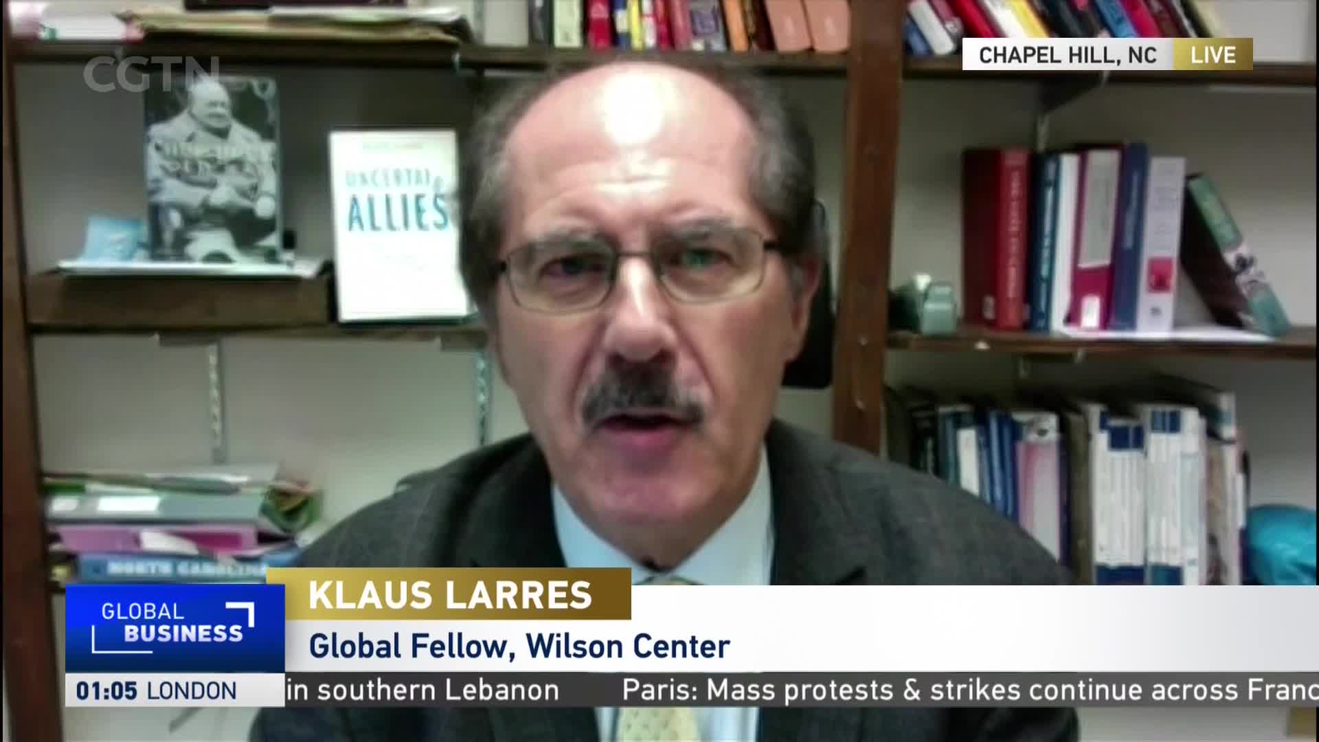 A man with a mustache and glasses speaks directly into the camera, framed by bookshelves and a CGTN logo. A chyron identifies him as Klaus Larres, a Global Fellow, as the broadcast originates from Chapel Hill, North Carolina.
