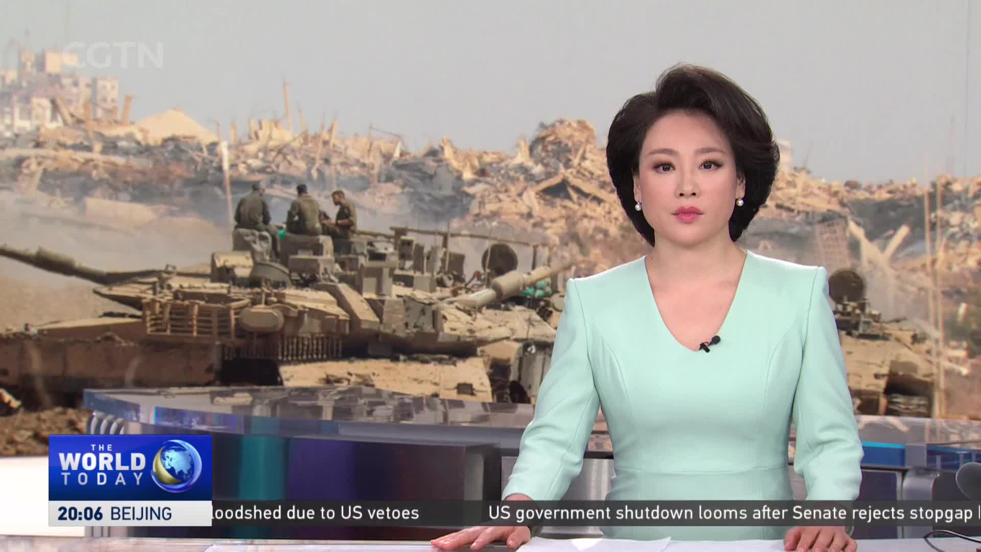 A woman in a mint-green jacket stands at a desk, speaking to the camera. Behind her, a tank and soldiers are visible against a backdrop of rubble, as seen on CGTN.
