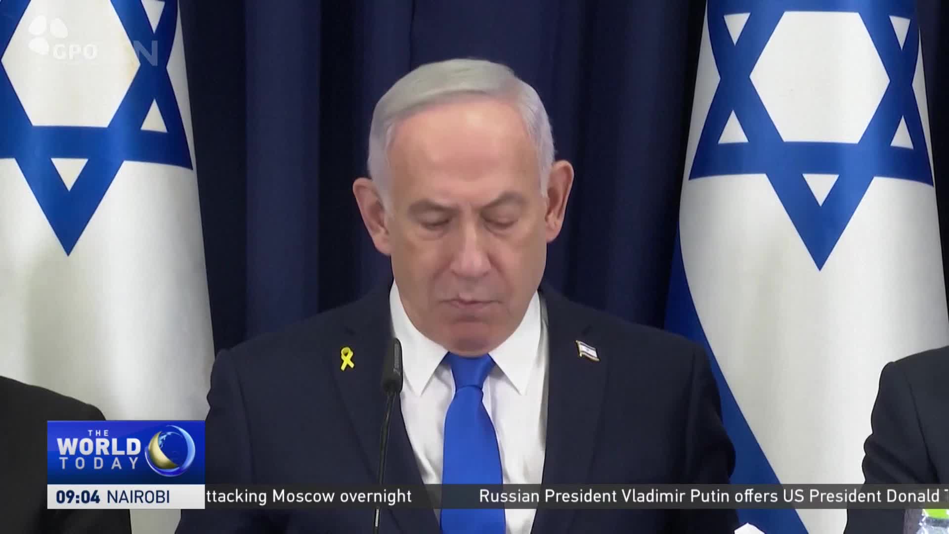 A man in a blue tie and dark suit stands at a podium, speaking. Behind him, two Israeli flags are visible on either side, and a CGTN news graphic is in the lower left corner.
