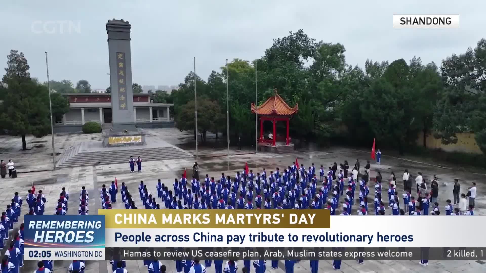 A large group of people in blue uniforms stand at attention, facing a tall monument. Two figures in the foreground carry a red flag, as CGTN broadcasts the event from Shandong, China.
