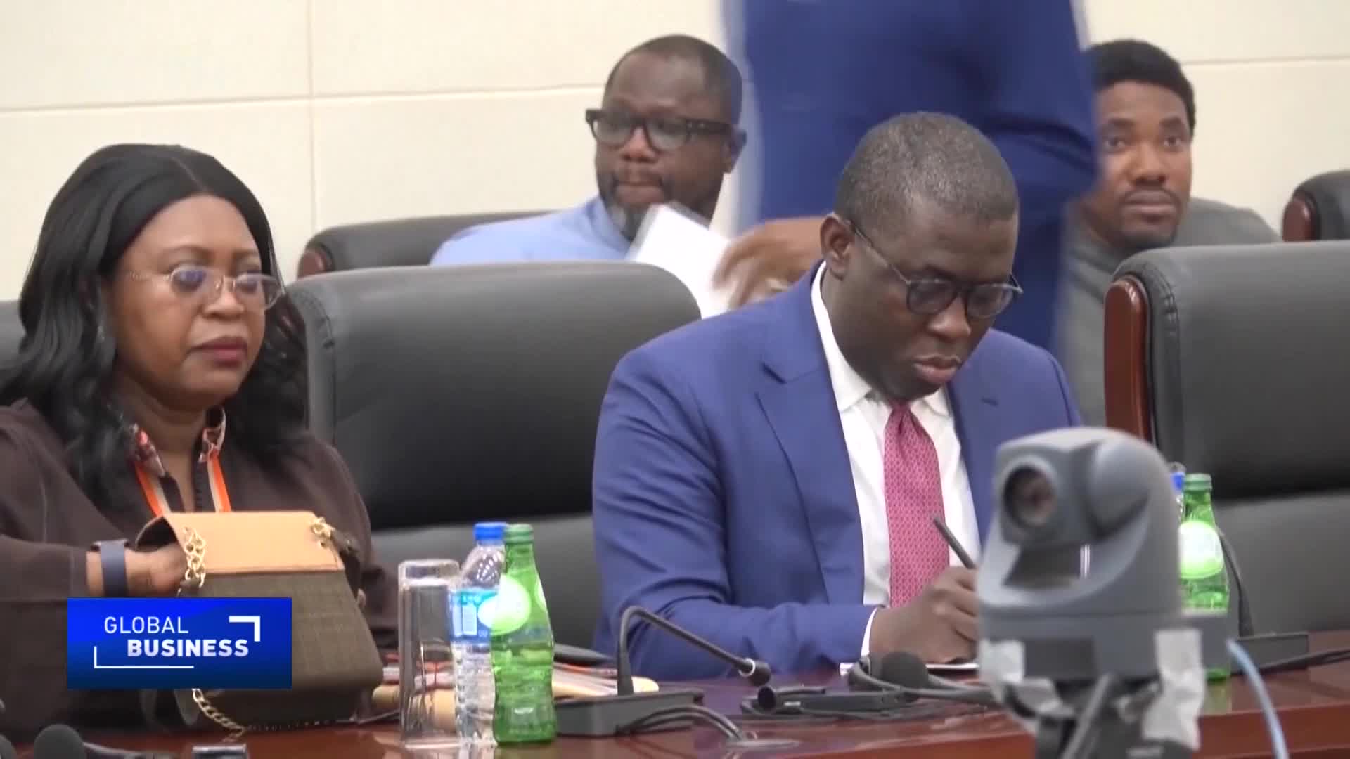 A man in a blue suit, glasses, and a red tie is focused on writing, his pen moving across a notepad.  Others are seated around a table, and a camera sits in the foreground, likely capturing the scene for CGTN.
