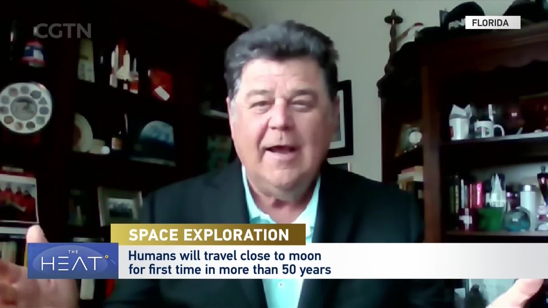 A man in a black suit jacket over a light blue shirt speaks, with a CGTN graphic overlaying the bottom of the screen. Behind him, shelves are filled with books and various decorative items, including what appear to be model rockets.