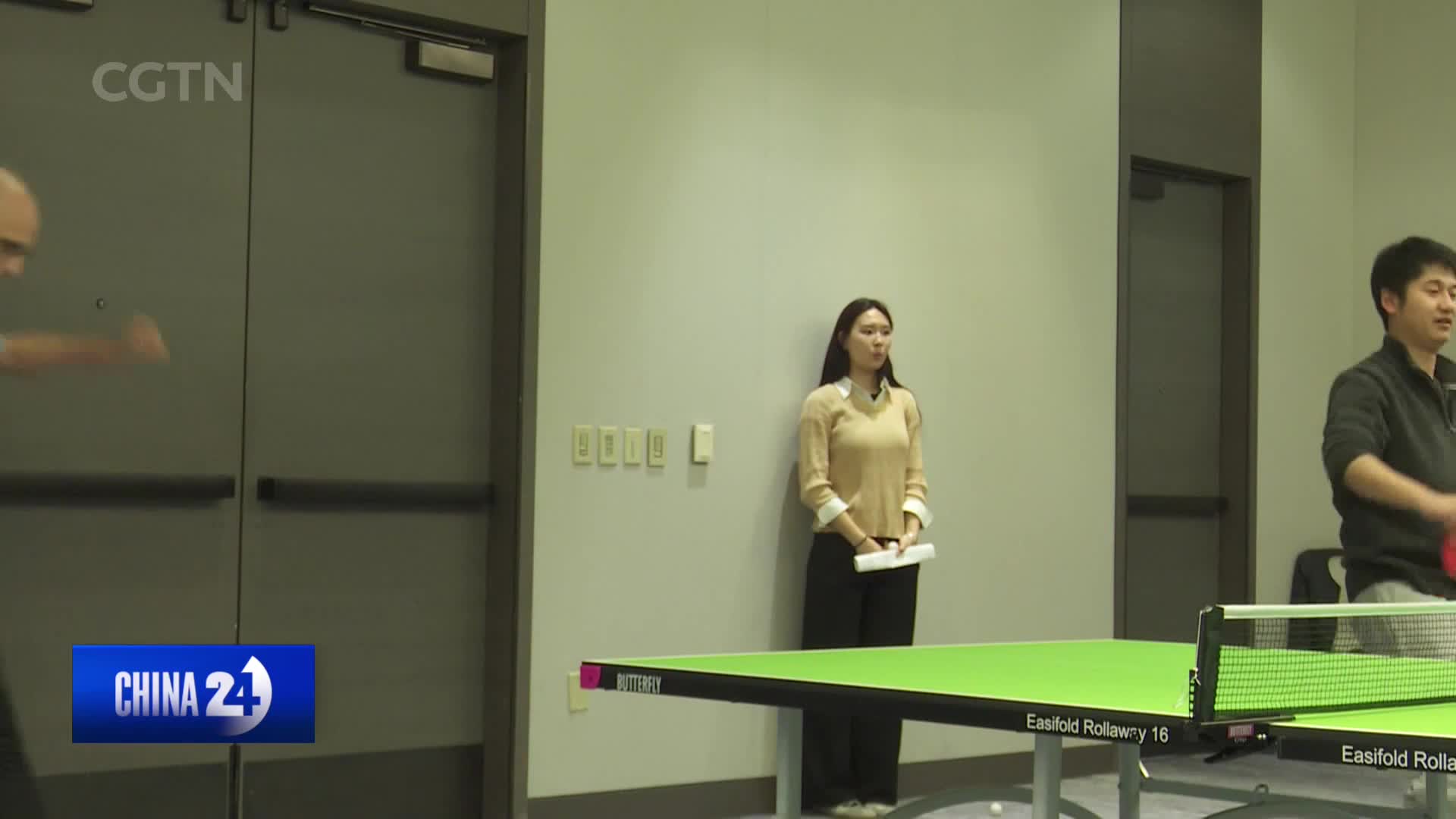 A man on the right swings a red paddle, his arm a blur as he hits the ball. A woman stands watching, holding a paper, while another man's arm is visible on the left, mid-swing.