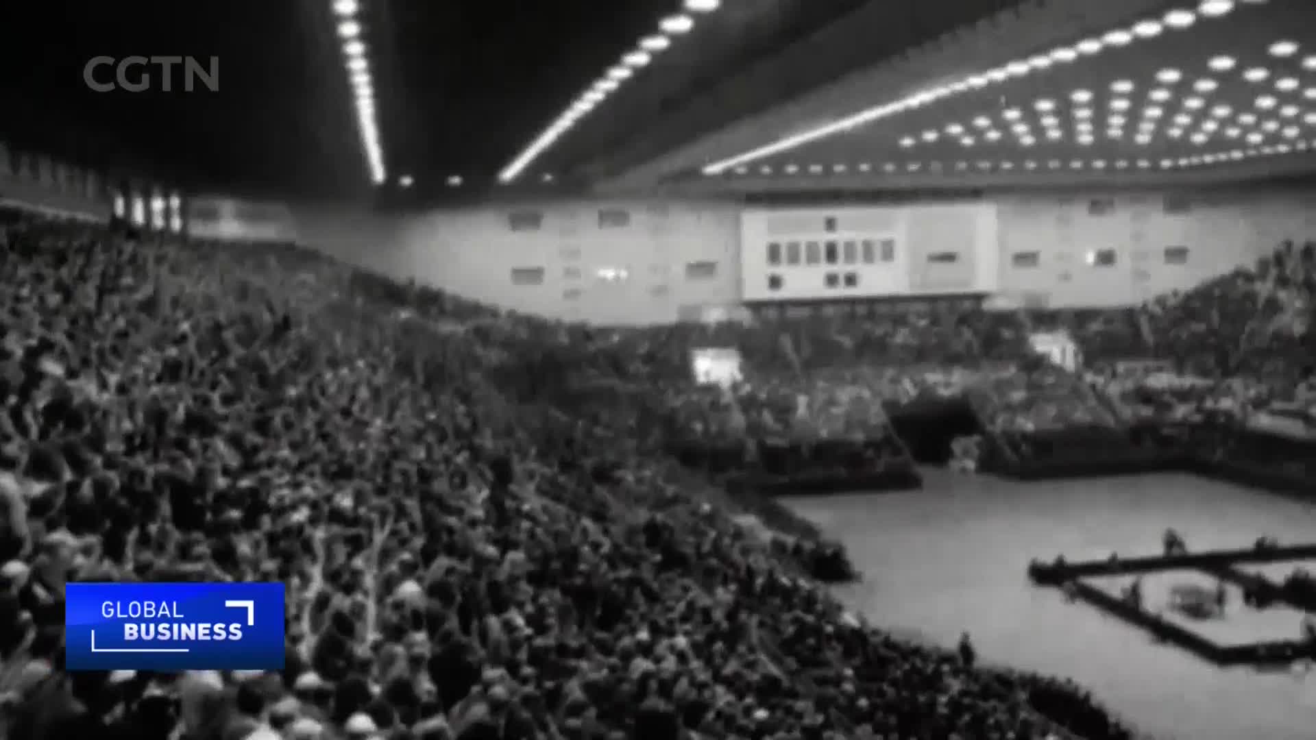 A vast crowd fills the tiered seating of a large indoor arena in China. The spectators are looking towards a central performance area, where figures are gathered on a raised platform.