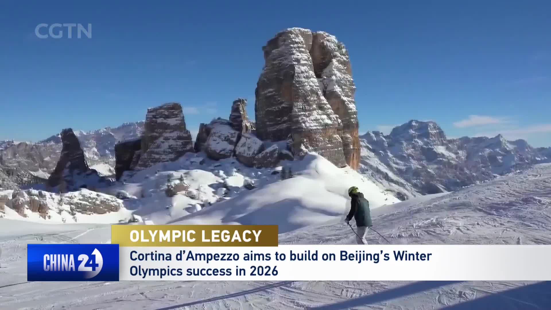 A lone skier carves down a pristine, snow-covered slope. Towering, rocky peaks, dusted with snow, rise majestically against a clear blue sky.