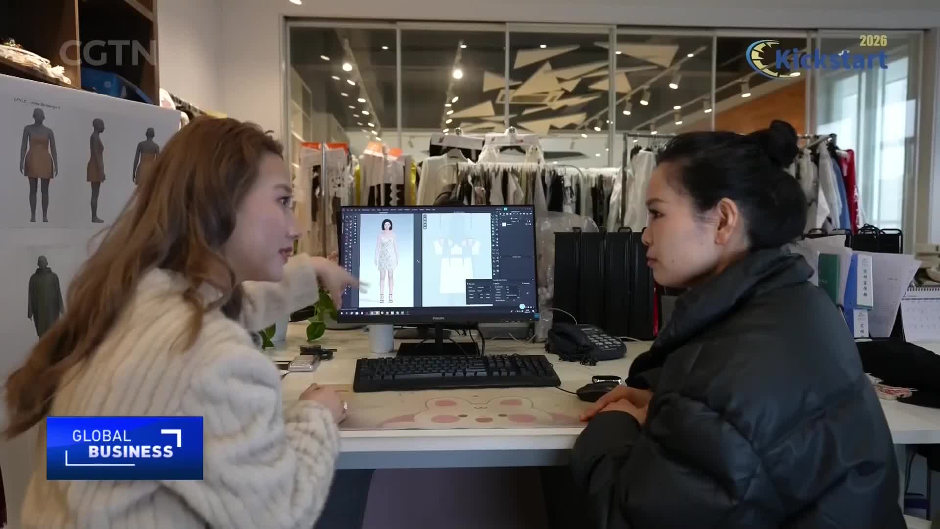 A woman points at a 3D rendering of a dress on a computer screen, discussing design details with her colleague. Behind them, racks of clothing hang, hinting at the busy fashion studio environment.