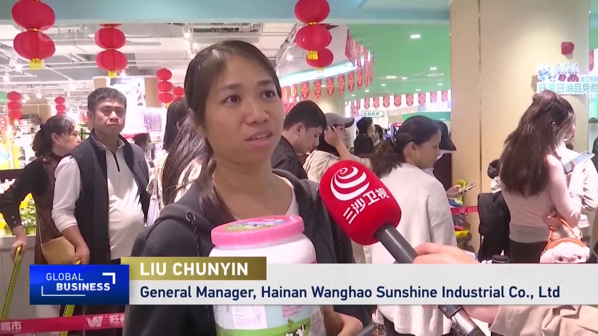 A woman holds a large pink and white container, speaking into a red microphone with Chinese characters. Behind her, shoppers move through a brightly lit store adorned with red lanterns. A woman holds a large pink and white container, speaking into a red microphone with Chinese characters. Behind her, shoppers move through a brightly lit store adorned with red lanterns.