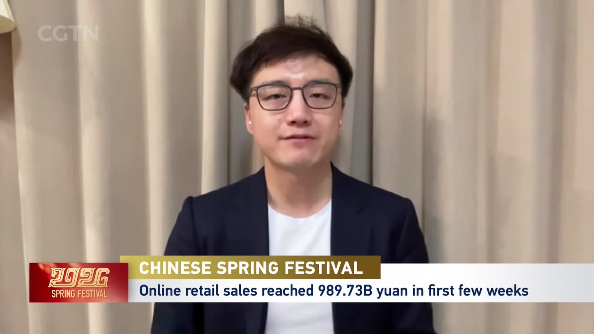 A man in a dark suit and glasses speaks directly to the camera, with a CGTN graphic overlay indicating "Chinese Spring Festival" and "Online retail sales reached 989.73B yuan in first few weeks." The background is a soft, beige curtain. A man in a dark suit and glasses speaks directly to the camera, with a CGTN graphic overlay indicating "Chinese Spring Festival" and "Online retail sales reached 989.73B yuan in first few weeks." The background is a soft, beige curtain.