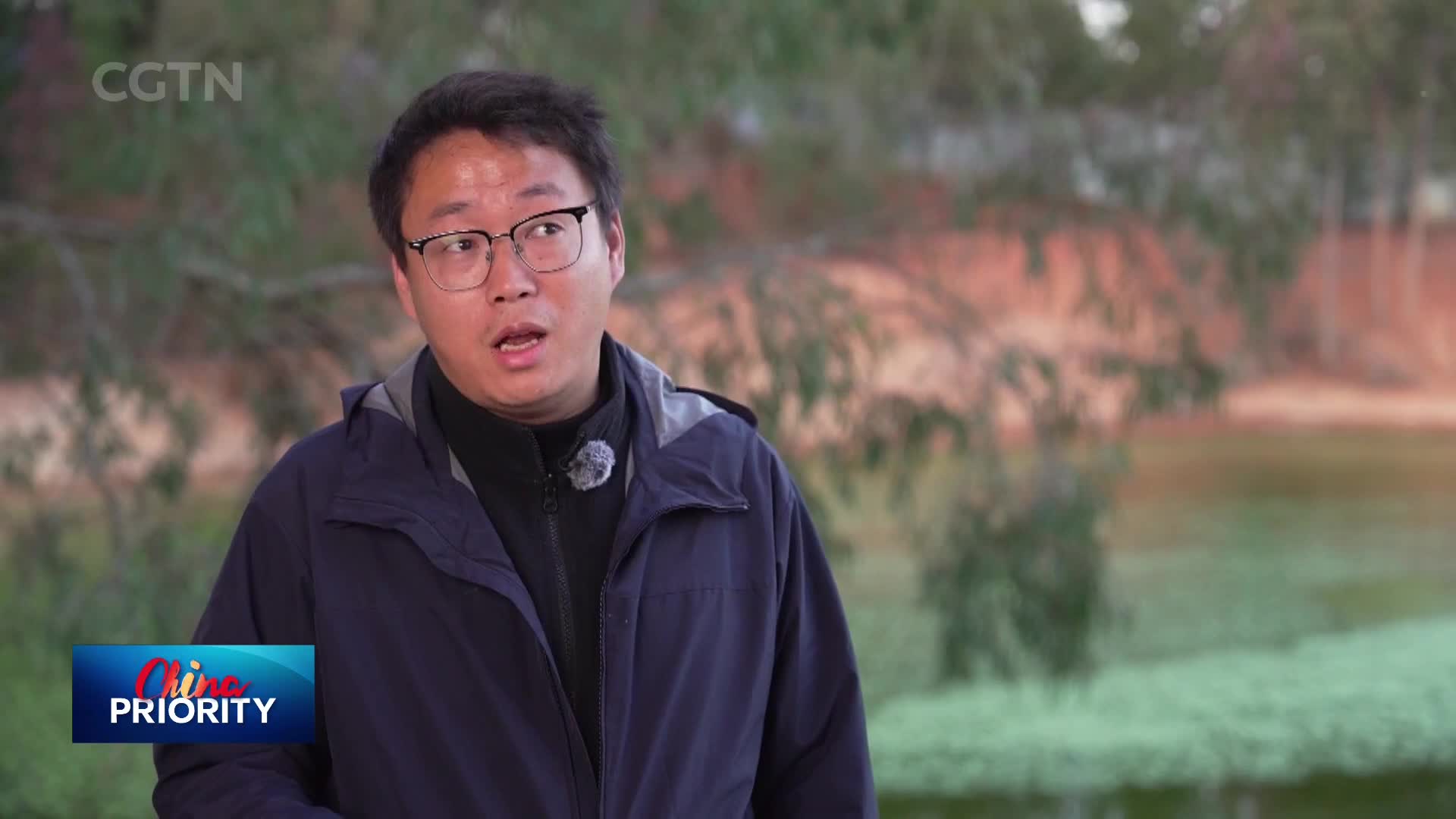 A man in a dark jacket speaks, his mouth slightly open. Behind him, trees and water create a soft, natural backdrop. A man in a dark jacket speaks, his mouth slightly open. Behind him, trees and water create a soft, natural backdrop.