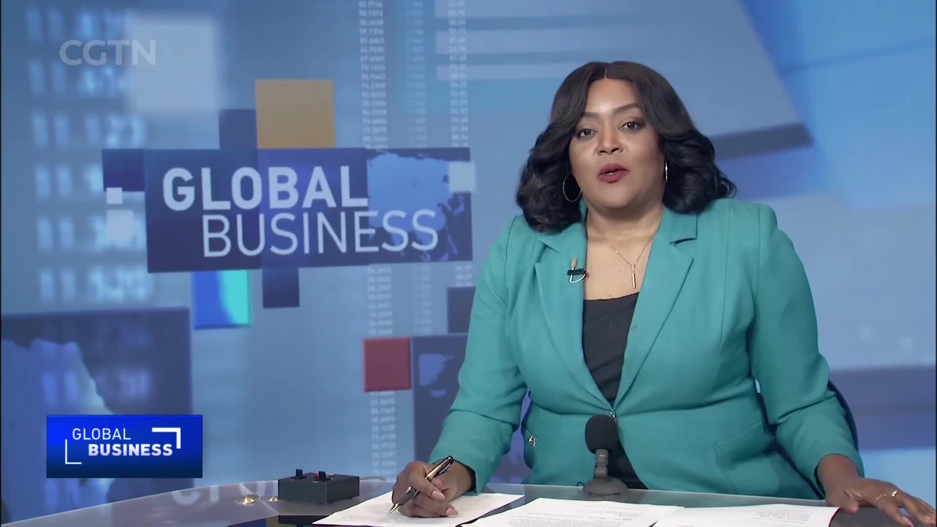 A woman in a teal blazer speaks into a microphone, her hand resting on papers. Behind her, the CGTN "Global Business" logo appears on a screen showing financial data.