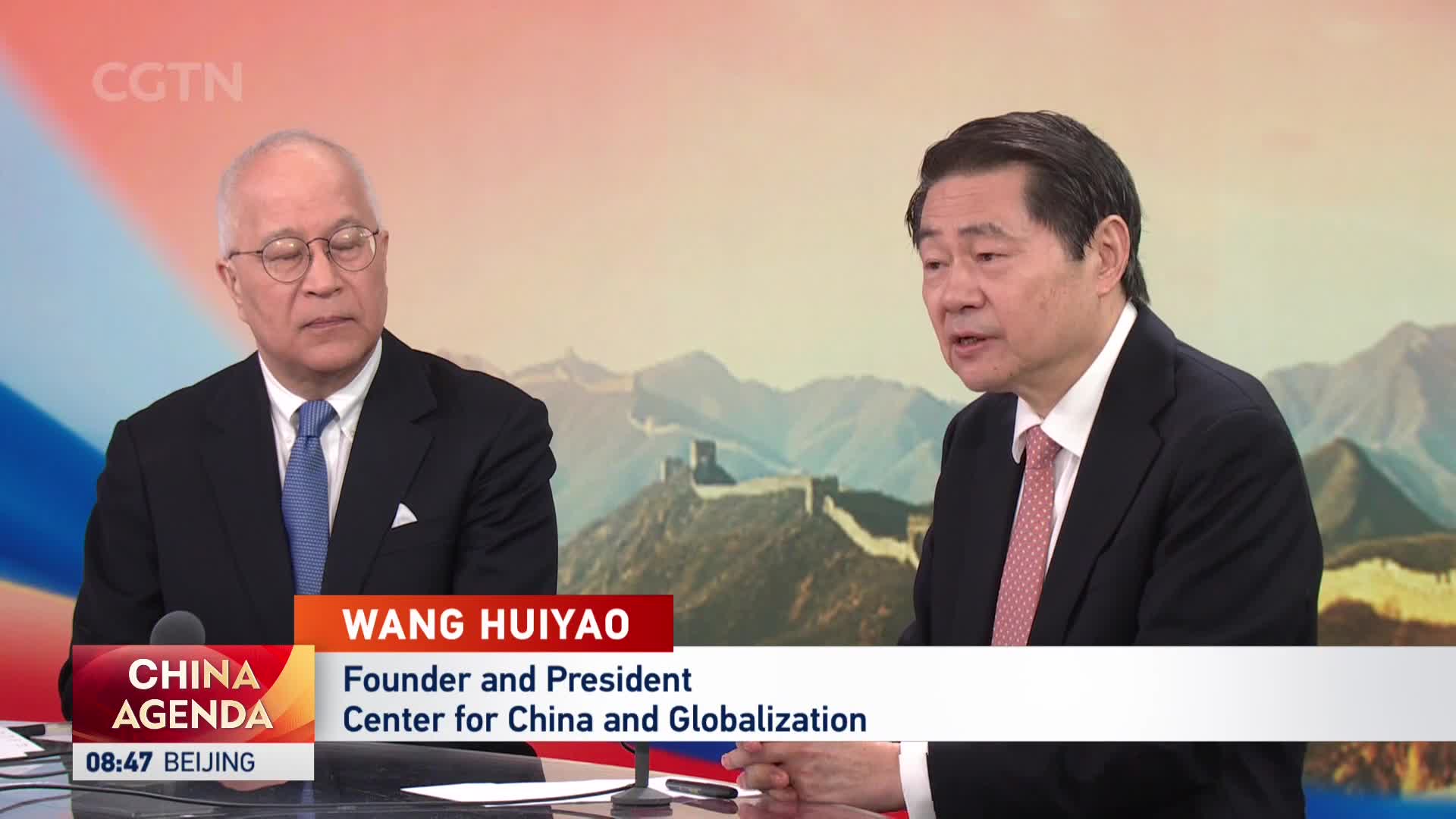 Two men sit at a table, one with glasses and a blue tie, the other in a dark suit with a patterned tie. A graphic identifies the man on the right as Wang Huiyao, Founder and President of the Center for China and Globalization, as the CGTN program "China Agenda" plays in the background.